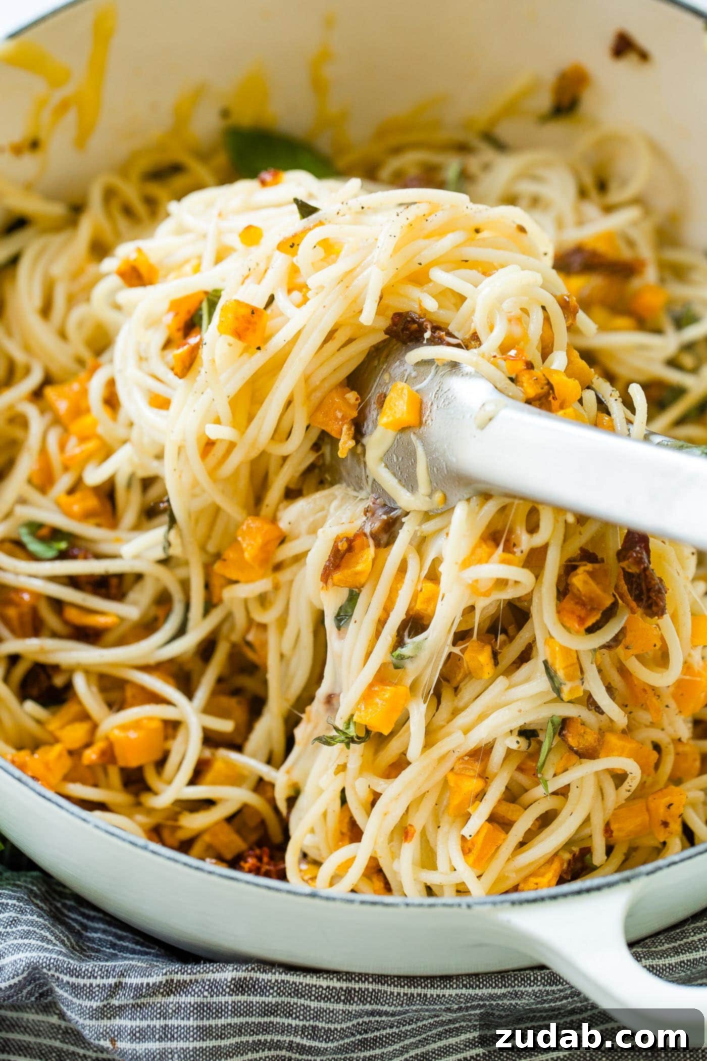 25 Minute Vegetarian Butternut Squash Pasta A photo of a pot full of butternut squash pasta being dished up with tongs. The steam rises gently, highlighting the fresh basil, sun-dried tomatoes, and golden squash mixed with spaghetti, ready for a delicious meal.