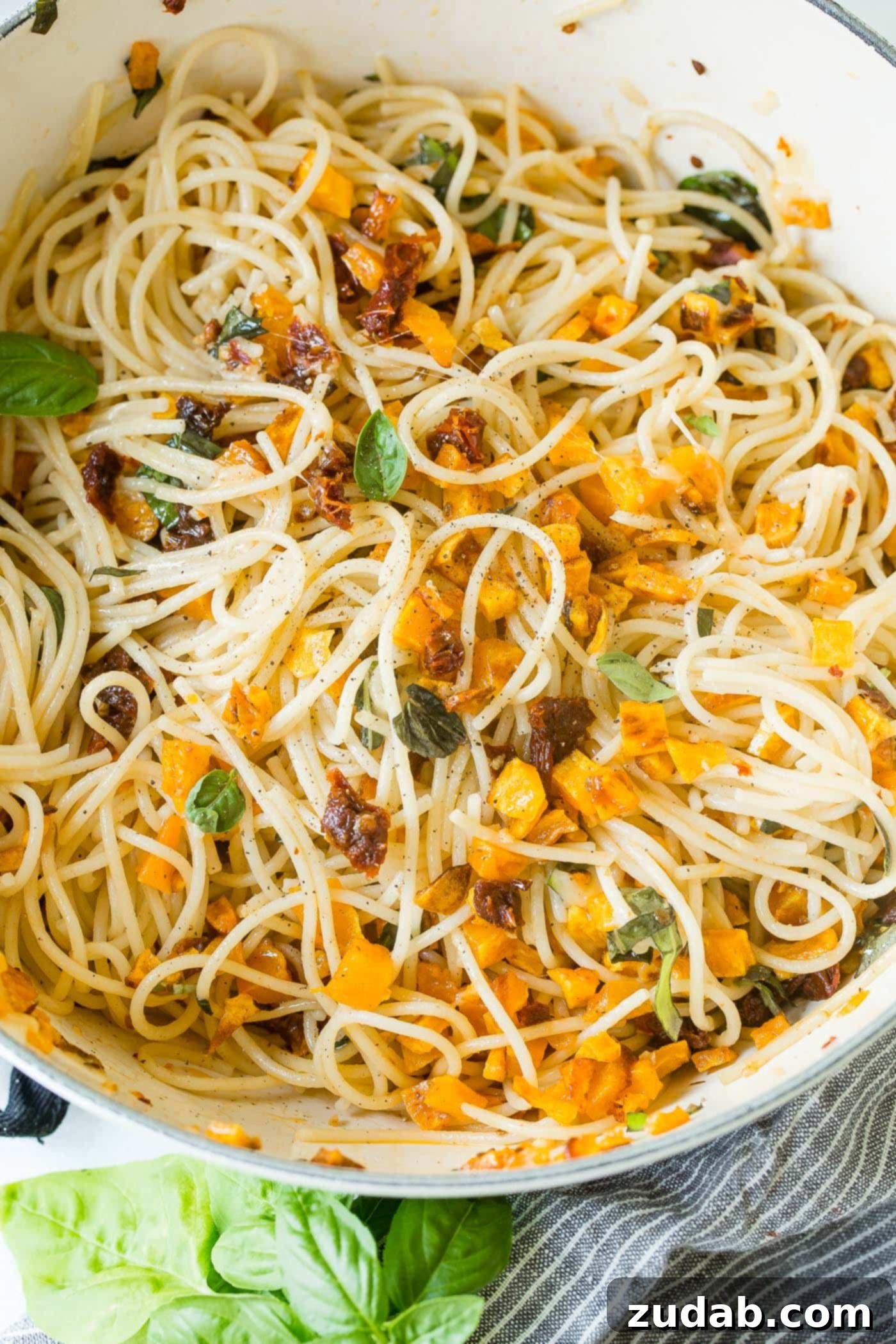 25 Minute Vegetarian Butternut Squash Pasta A vibrant close-up photo of a large pot of butternut squash pasta, overflowing with cooked spaghetti, roasted golden butternut squash cubes, bright red sun-dried tomatoes, and fresh green basil leaves. The dish is rich and ready to be enjoyed.