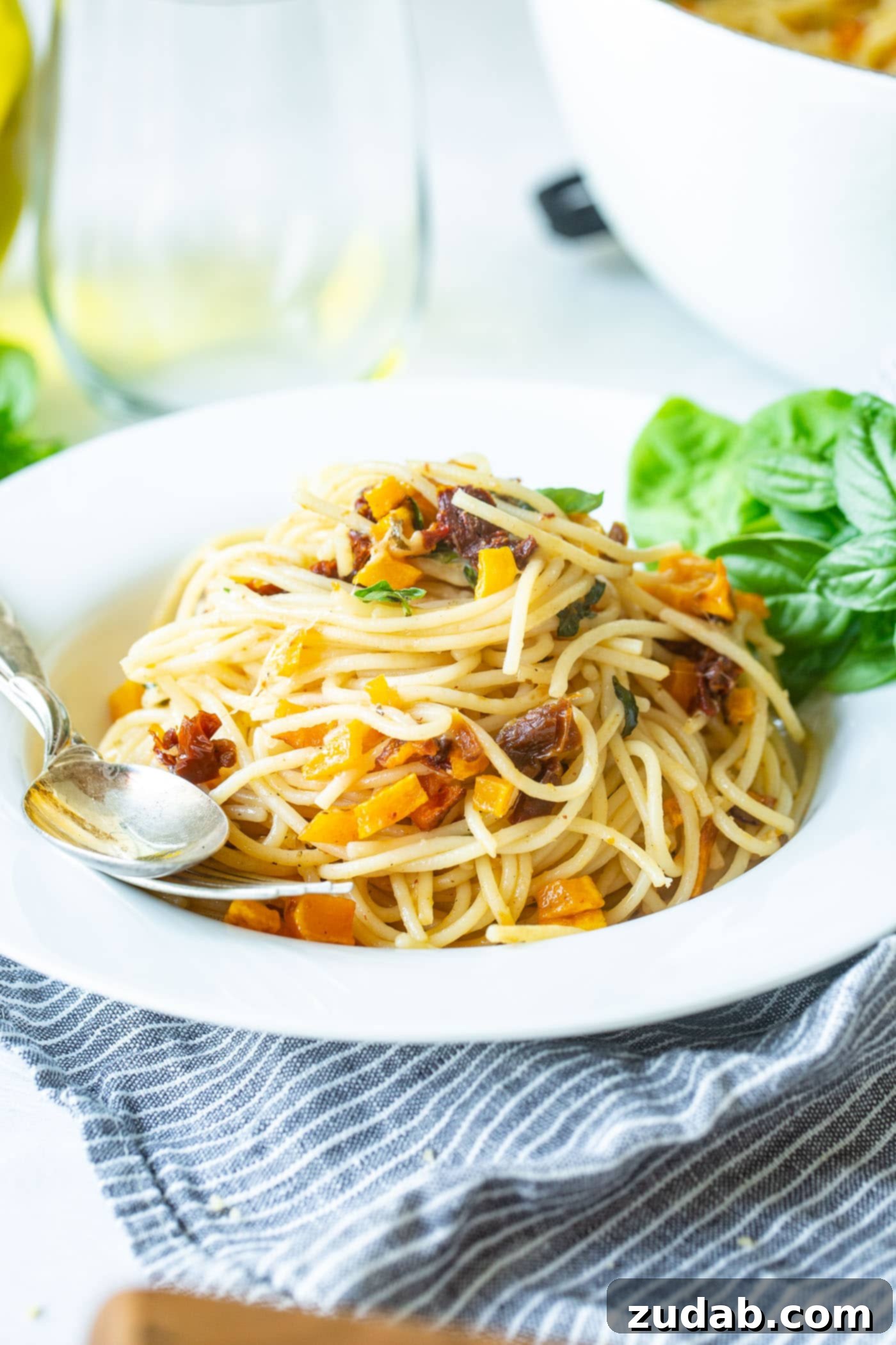 25 Minute Vegetarian Butternut Squash Pasta A vibrant serving of butternut squash pasta in a white bowl with a fork and spoon on the side. The dish features golden squash, bright sun-dried tomatoes, fresh basil, and melted cheese, making for an inviting vegetarian meal.