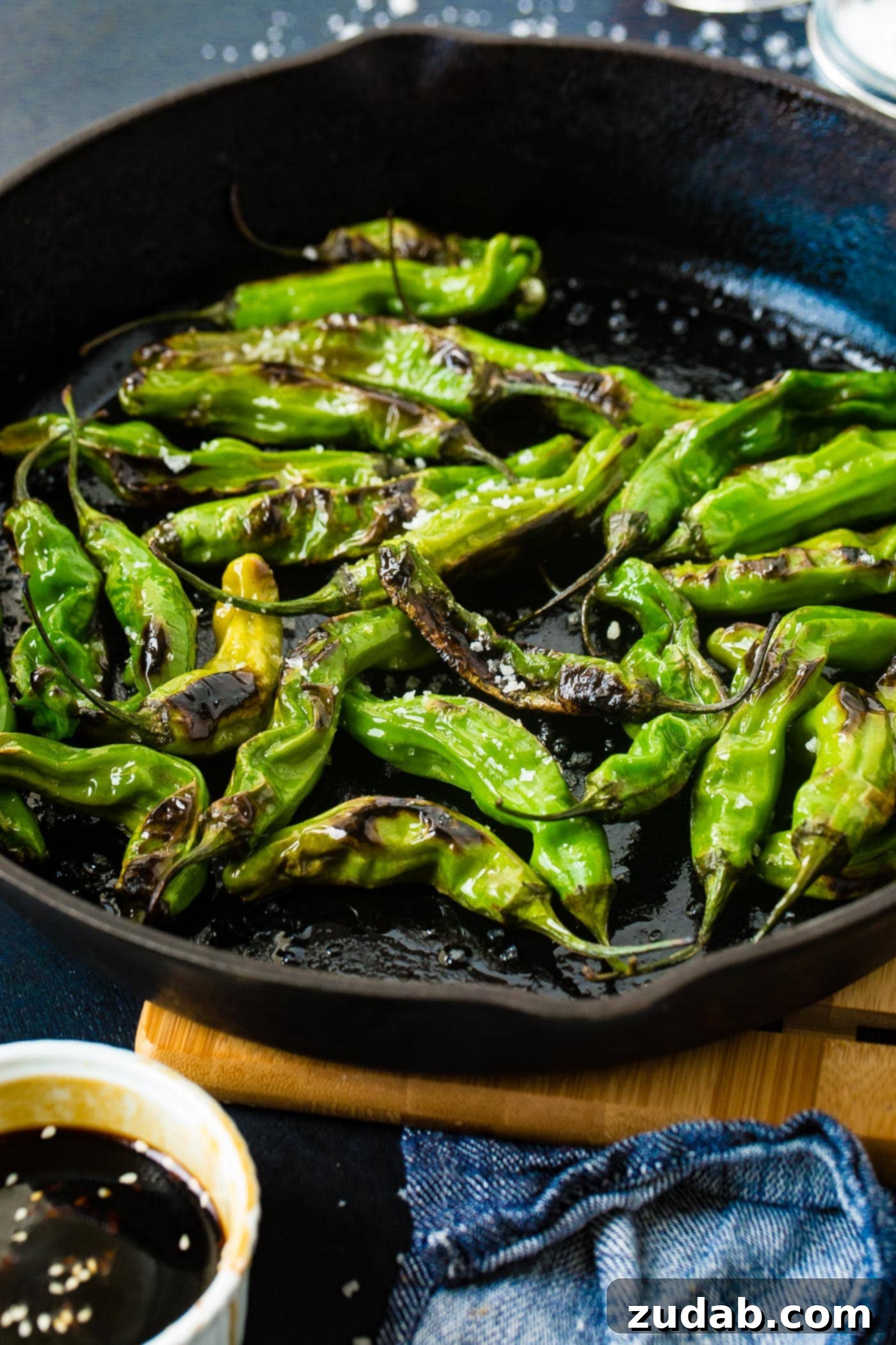 How to Get Perfectly Blistered Shishito Peppers 2 A black cast iron pan overflowing with bright green blistered shishito peppers, generously sprinkled with glistening flaked sea salt, creating an irresistible visual.