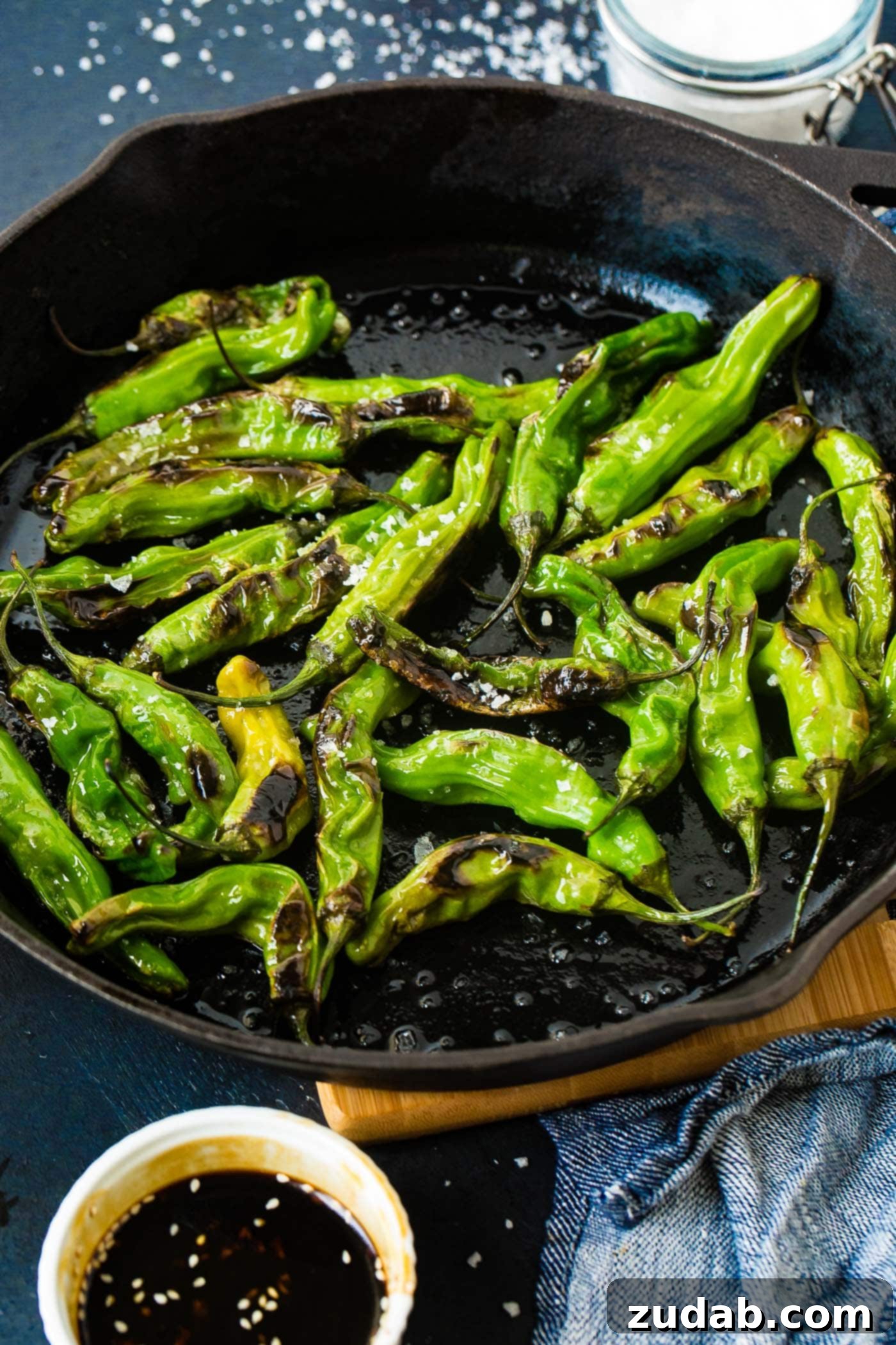 How to Get Perfectly Blistered Shishito Peppers 4 A third captivating shot of the black cast iron pan, laden with bright green blistered shishito peppers and a generous scattering of flaked sea salt, creating an irresistible snack.