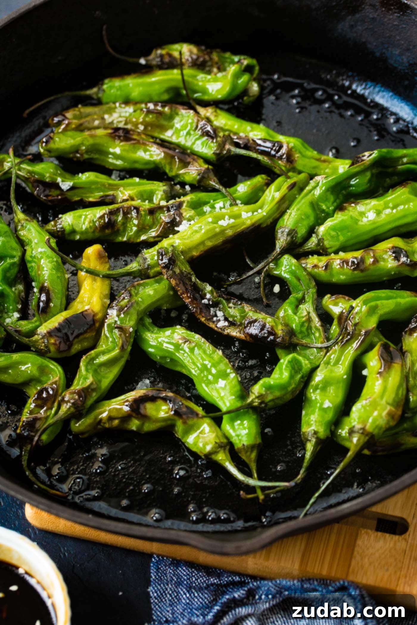 How to Get Perfectly Blistered Shishito Peppers 5 A final close-up shot of the black cast iron pan filled with bright green blistered shishito peppers, beautifully seasoned with flaked sea salt, emphasizing their vibrant color and texture.