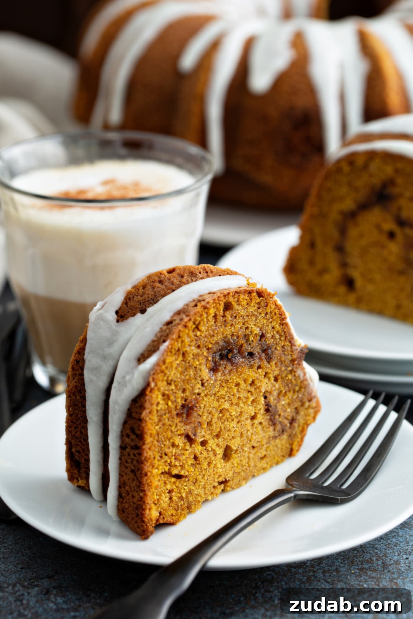 Sour Cream Cinnamon Swirl Pumpkin Bundt Cake A perfectly sliced piece of Sour Cream Cinnamon Swirl Pumpkin Bundt Cake on a white dessert plate, with a fork resting beside it. The cake features a beautiful white glaze drizzled over the top and a visible cinnamon swirl within its moist texture. In the background, another slice and the main bundt cake on a decorative platter complete the inviting scene.