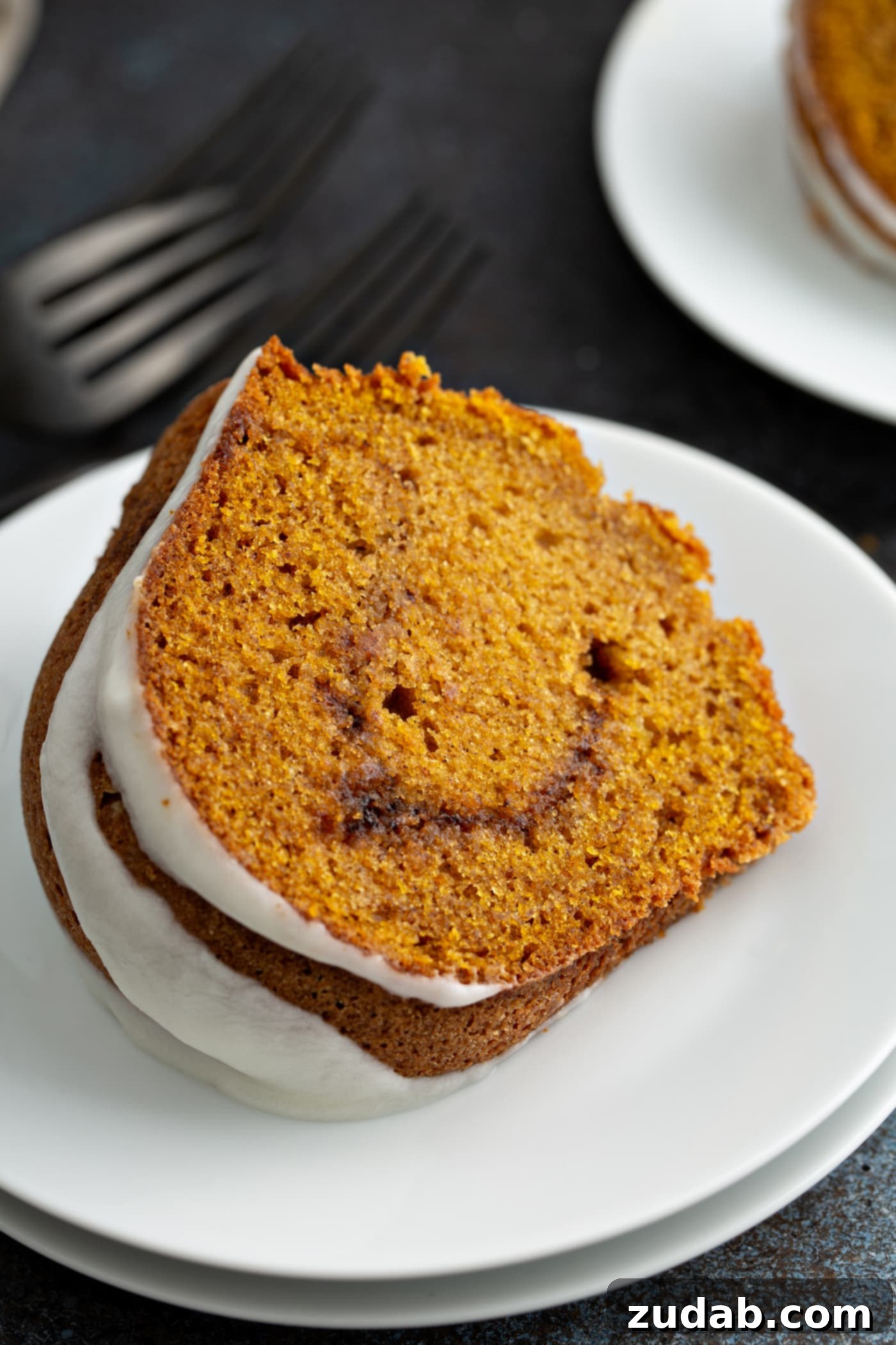 Sour Cream Cinnamon Swirl Pumpkin Bundt Cake A single, delectable slice of Sour Cream Cinnamon Swirl Pumpkin Cake rests on a white dessert plate. The cake showcases a beautiful drizzle of glaze over its top and a clear view of the rich cinnamon swirl within its layers. A fork lies gracefully beside the plate, with another dessert plate subtly in the background.