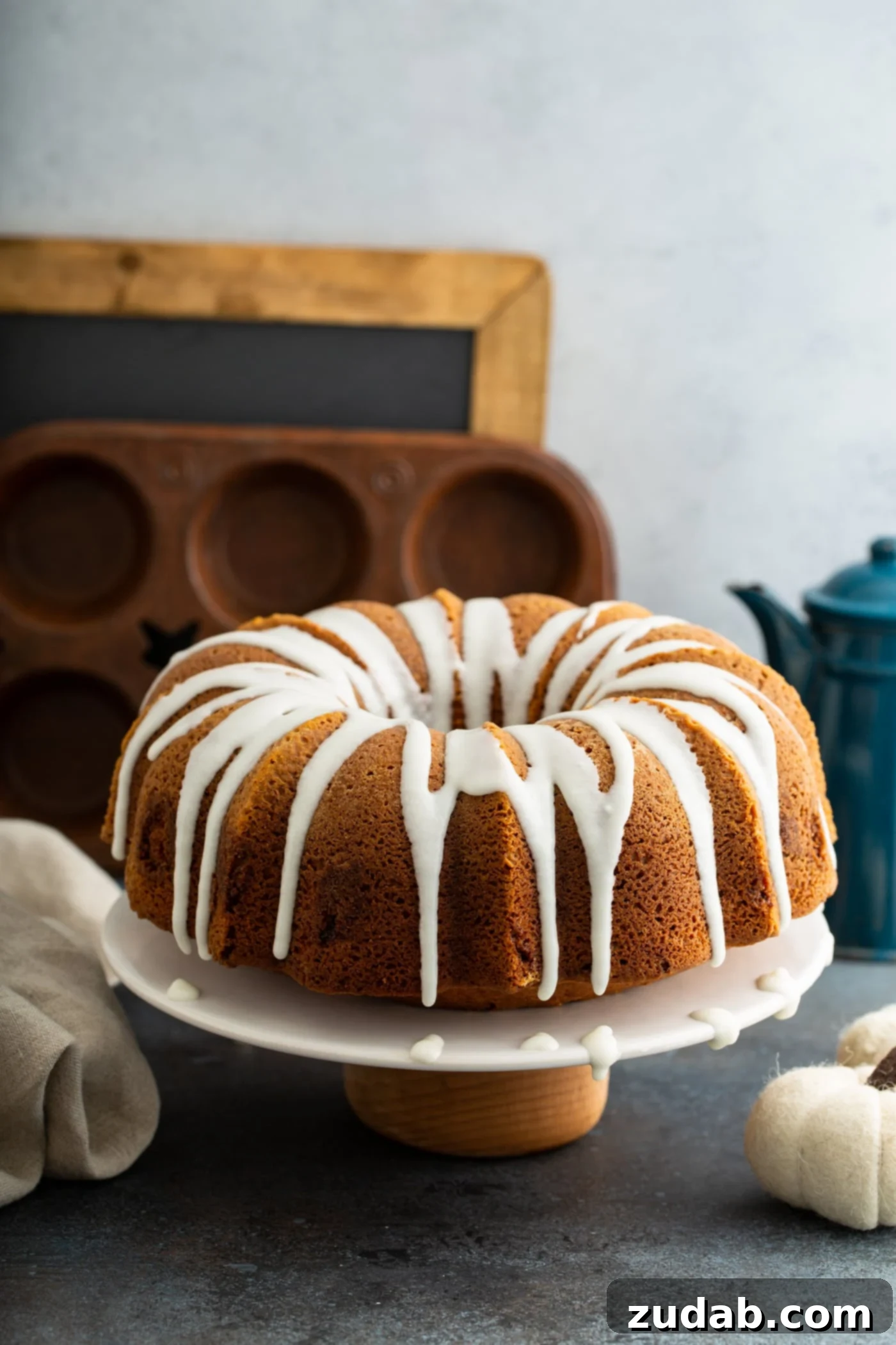 Sour Cream Cinnamon Swirl Pumpkin Bundt Cake A beautifully presented Sour Cream Cinnamon Swirl Pumpkin Bundt Cake, generously drizzled with white glaze, sits on a pristine white cake plate. In the tastefully blurred background, a muffin tin, a rustic chalk board, and a classic coffee pot create a warm and inviting kitchen ambiance.