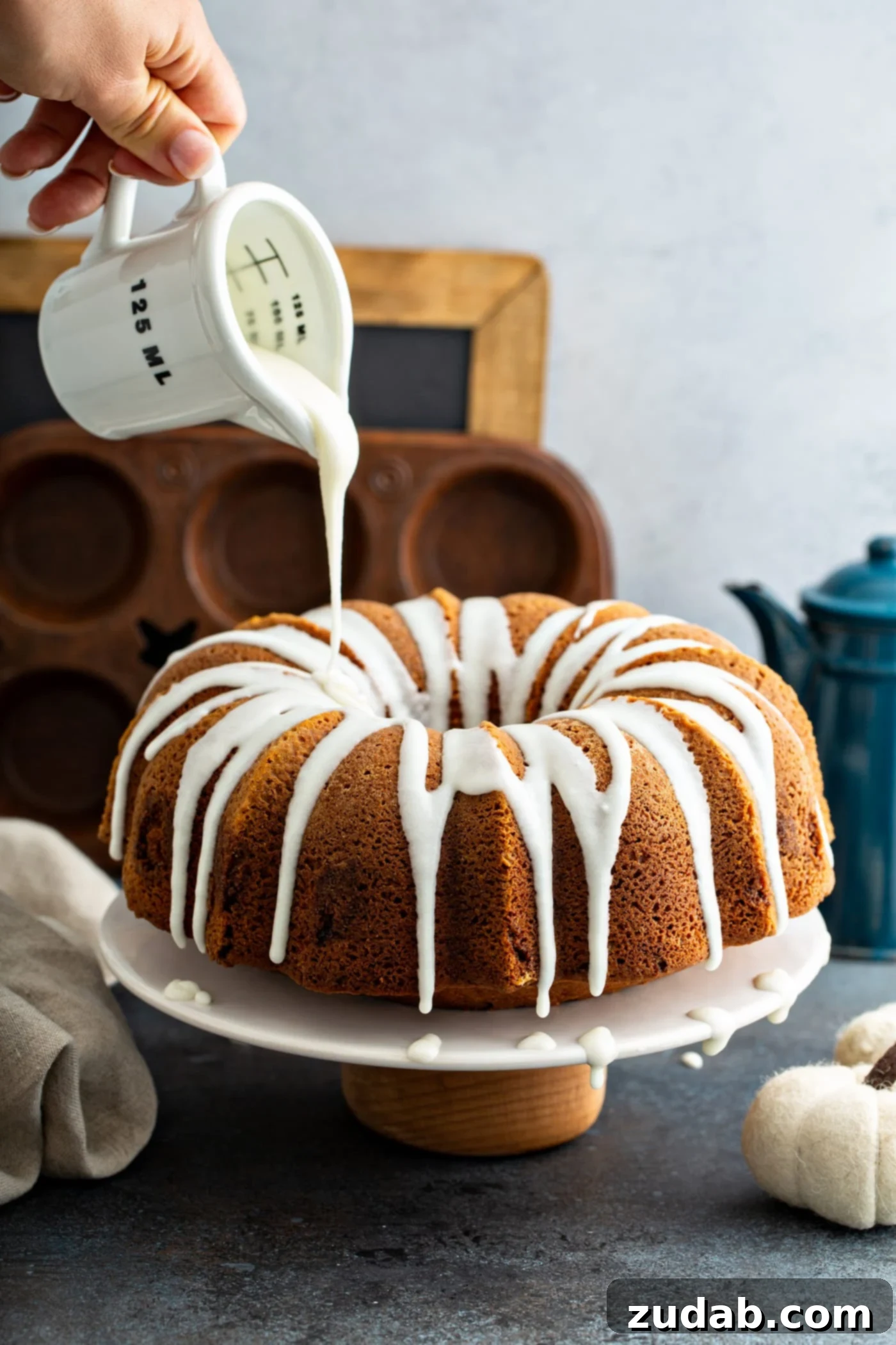 Sour Cream Cinnamon Swirl Pumpkin Bundt Cake White glaze being artfully drizzled from a clear glass measuring cup over the top of a freshly baked Sour Cream Cinnamon Swirl Pumpkin Bundt Cake, creating an inviting visual. In the soft-focus background, a muffin tin, a rustic chalk board, and a vintage blue coffee pot hint at a cozy kitchen setting.