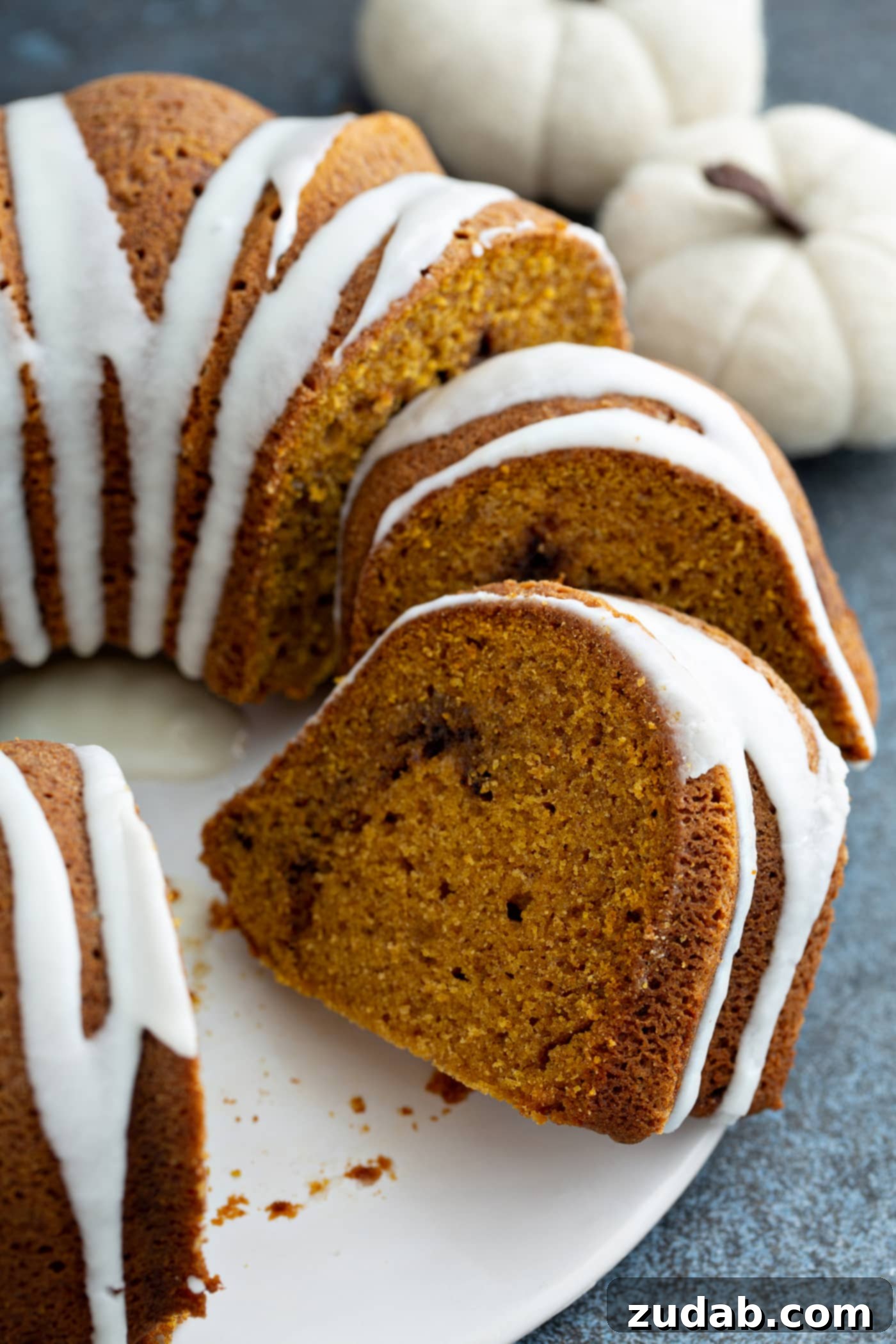 Sour Cream Cinnamon Swirl Pumpkin Bundt Cake A beautifully baked Sour Cream Cinnamon Swirl Pumpkin Bundt Cake presented on a white cake platter, with a few slices already cut. The cake is elegantly drizzled with a white frosting, highlighting its enticing shape and texture.