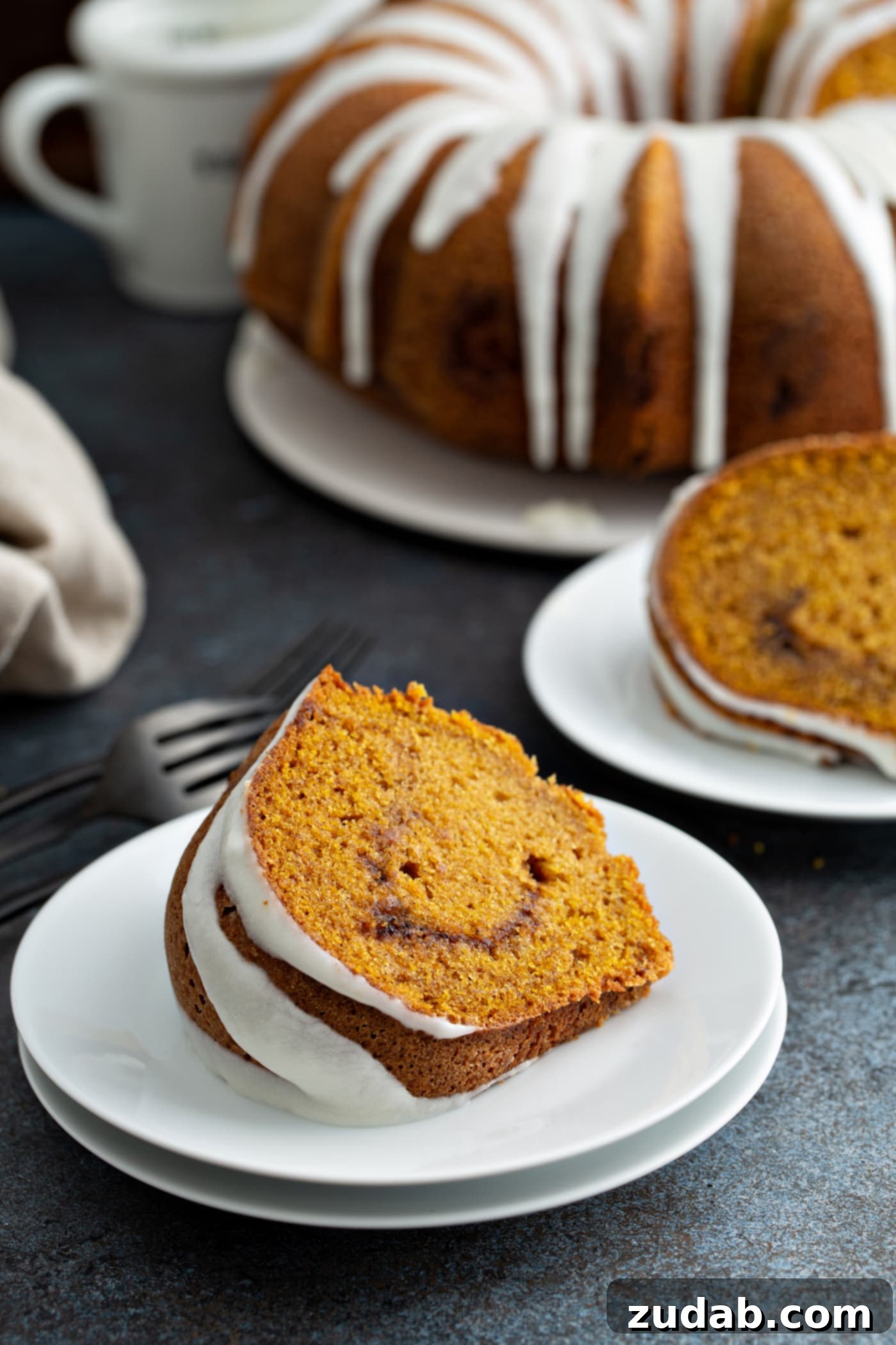 Sour Cream Cinnamon Swirl Pumpkin Bundt Cake Two enticing slices of Sour Cream Cinnamon Swirl Pumpkin Bundt Cake displayed on individual dessert plates. The distinct cinnamon swirl is clearly visible in the center of each slice, and the tops are delicately drizzled with a white glaze. In the background, the remaining bundt cake, also glazed, sits on a platter. A fork and napkin are neatly arranged beside one of the dessert plates.