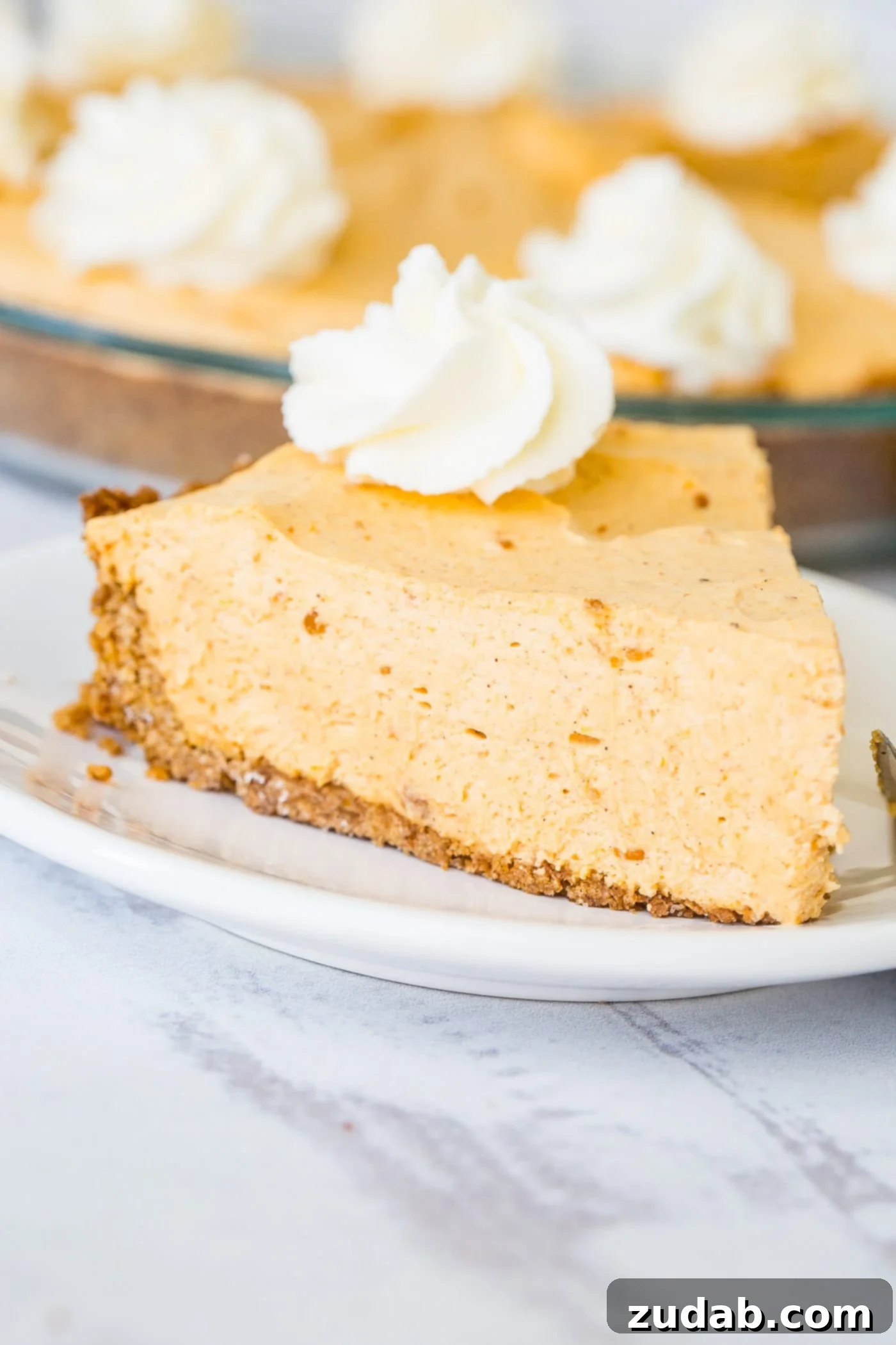 Marshmallow No Bake Pumpkin Pie A photo of a slice of no bake pumpkin pie on a white plate with a generous swirl of whipped cream on top, ready for a dessert spread.