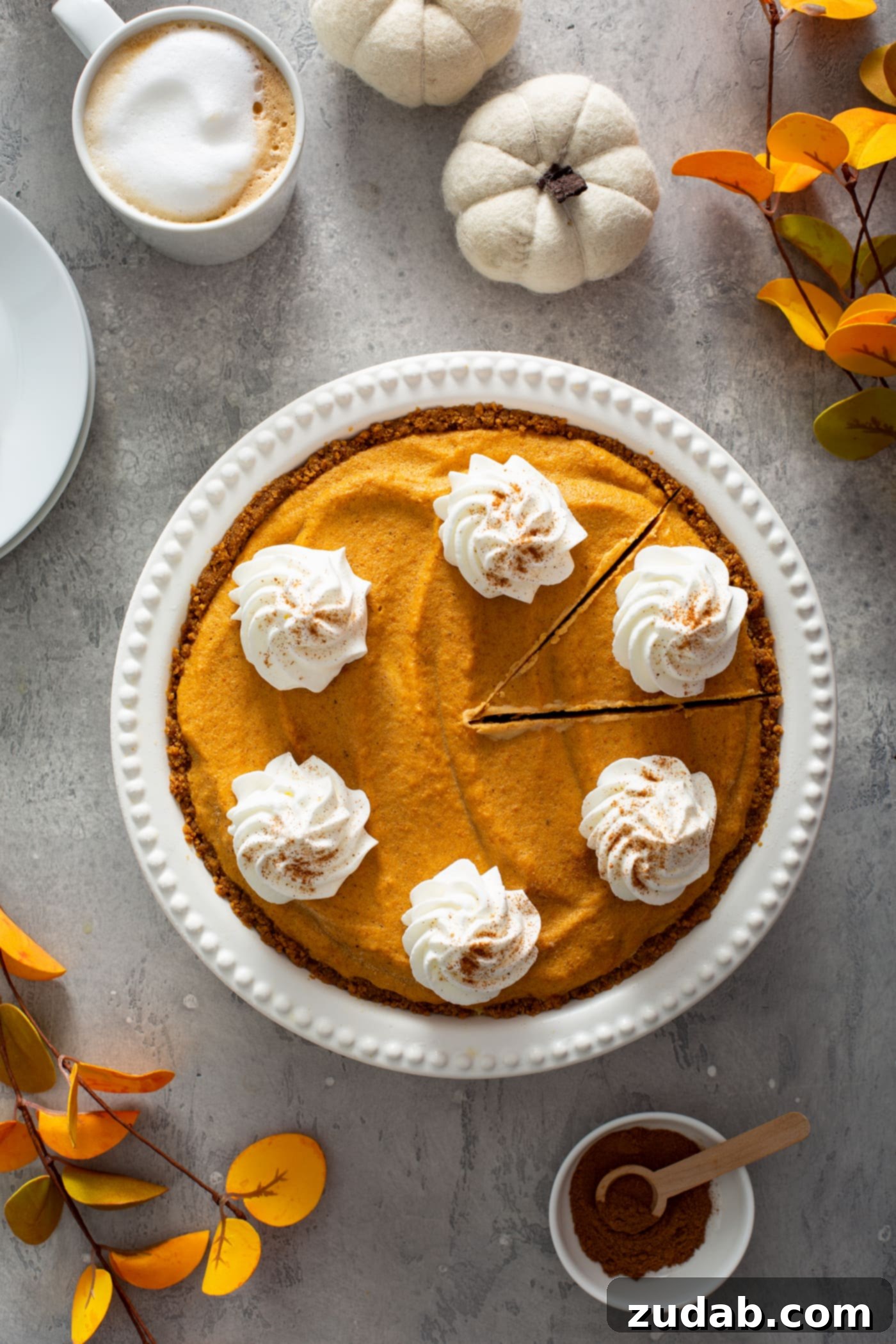 A whole pumpkin ice cream pie, beautifully presented in a white pie dish. The pie is artfully topped with generous dollops of whipped cream, each sprinkled with cinnamon. A slice has been cut but not yet removed from the pie, inviting indulgence. In the background, a dessert plate, a container of whipped cream, two tiny white pumpkins, vibrant fall-colored leaves, and a small bowl of cinnamon complete a picturesque and festive scene.