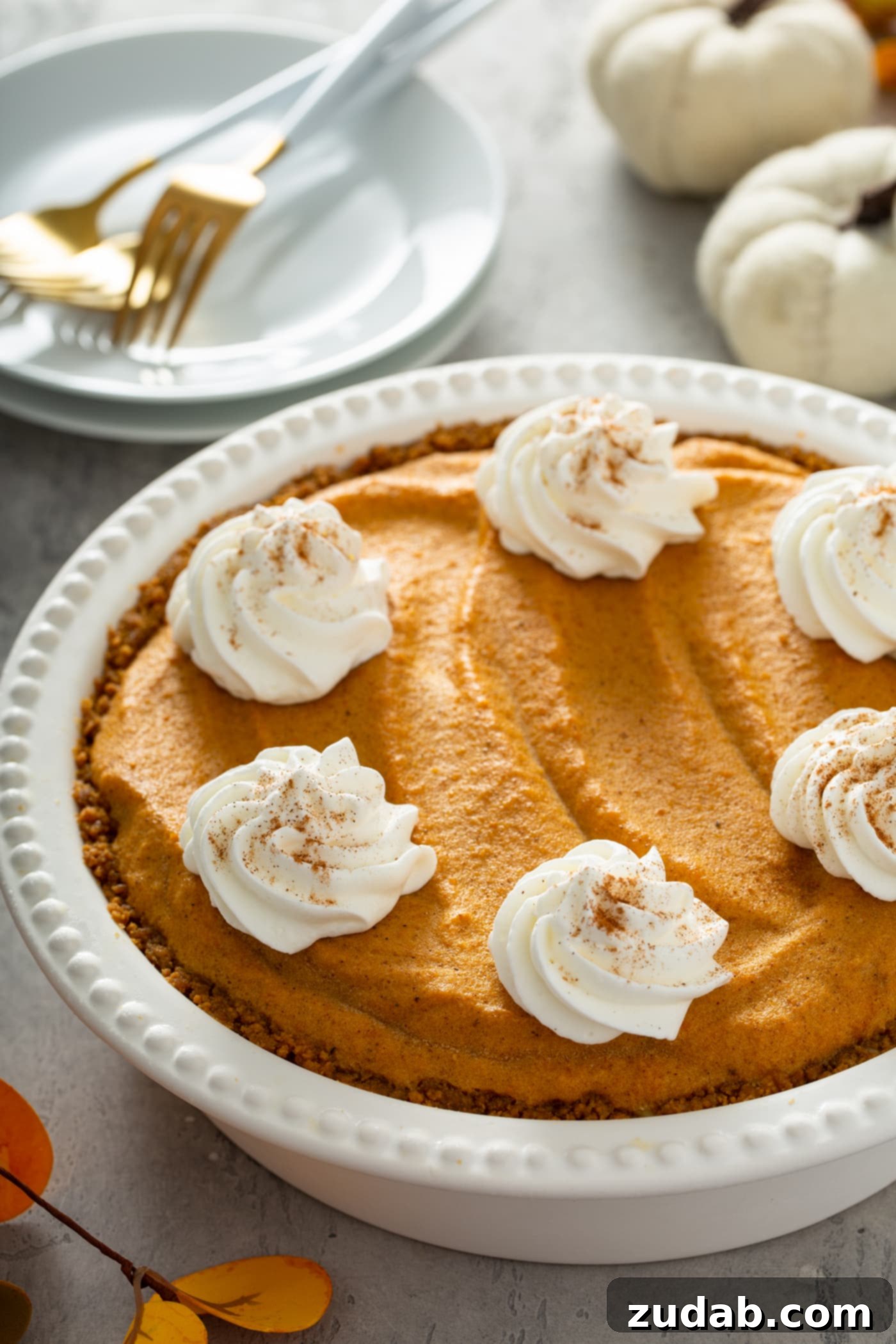 A pristine white pie dish cradles a whole pumpkin ice cream pie, generously adorned with dollops of whipped cream, each sprinkled with cinnamon. In the background, a neat stack of white dessert plates and two tiny white pumpkins complete a charming and festive tableau.