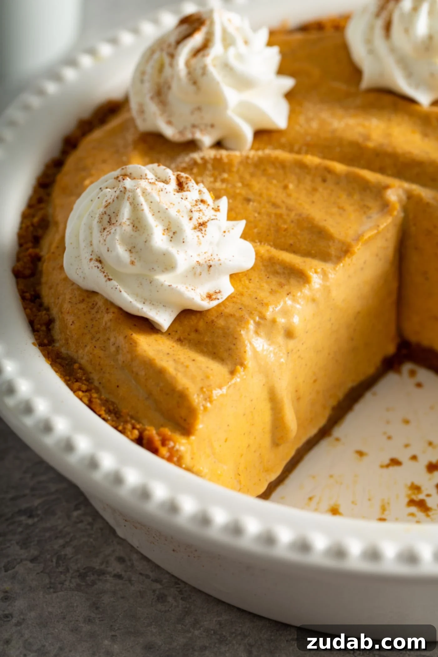 A pumpkin ice cream pie in a pristine white pie dish with a slice elegantly removed. The pie is generously topped with dollops of fluffy whipped cream, lightly dusted with cinnamon, inviting a delightful indulgence.