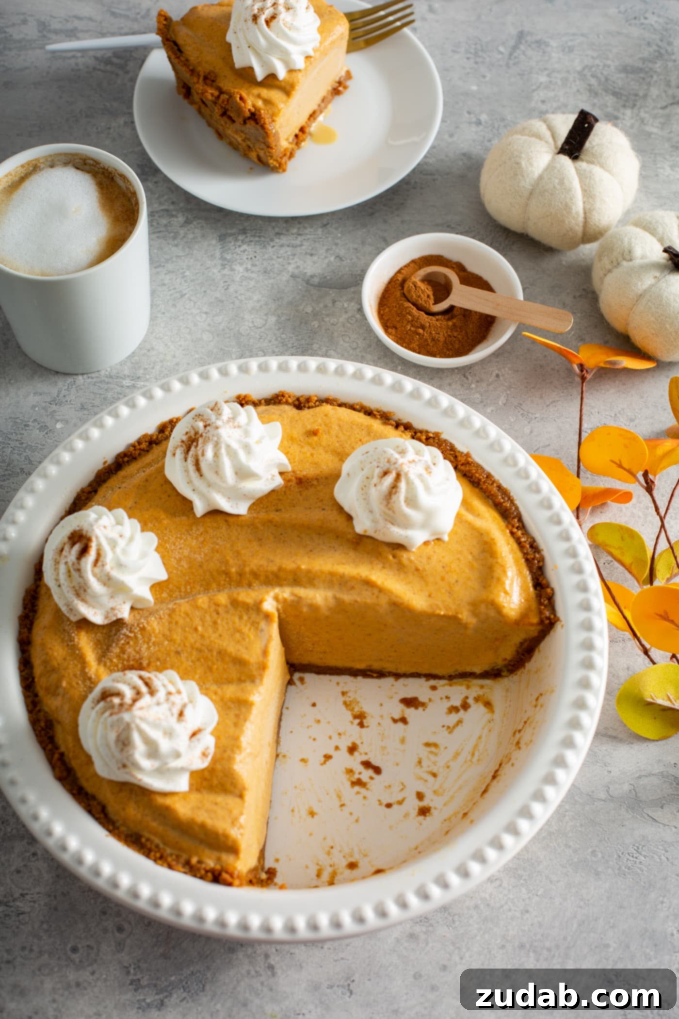 A white pie dish holds a tempting pumpkin ice cream pie, generously topped with dollops of whipped cream and a sprinkle of cinnamon. In the background, a white mug with a whipped cream-topped drink, a small dish of cinnamon with a wooden spoon, and two tiny white pumpkins complete a cozy fall scene.