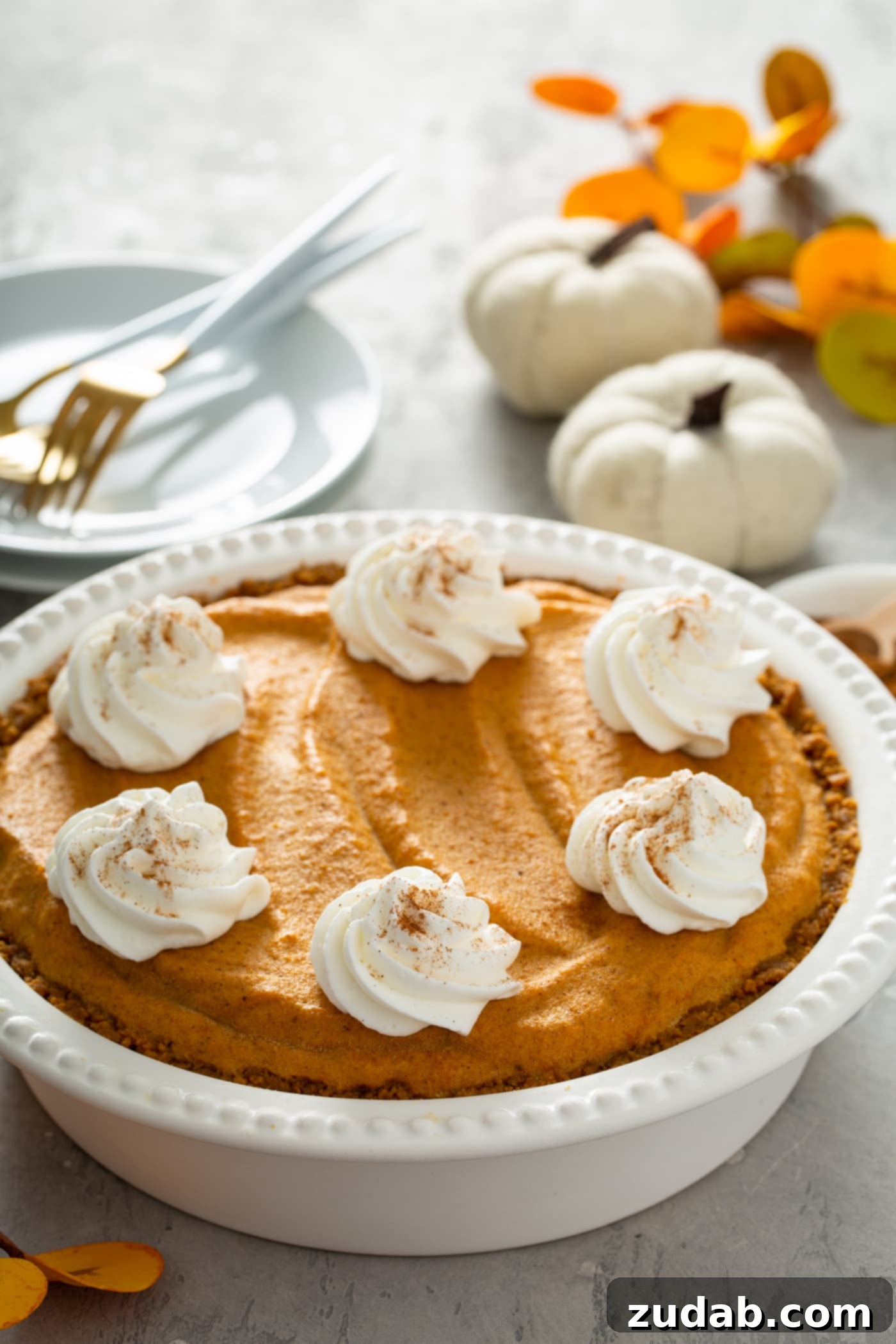 A visually stunning whole pumpkin ice cream pie, presented in a pristine white pie dish. The pie is beautifully topped with generous dollops of whipped cream, lightly dusted with cinnamon, creating an inviting contrast. Behind the pie, a stack of white dessert plates holds three forks, accompanied by two tiny white pumpkins and an arrangement of vibrant fall-colored leaves, setting a perfect autumnal table.