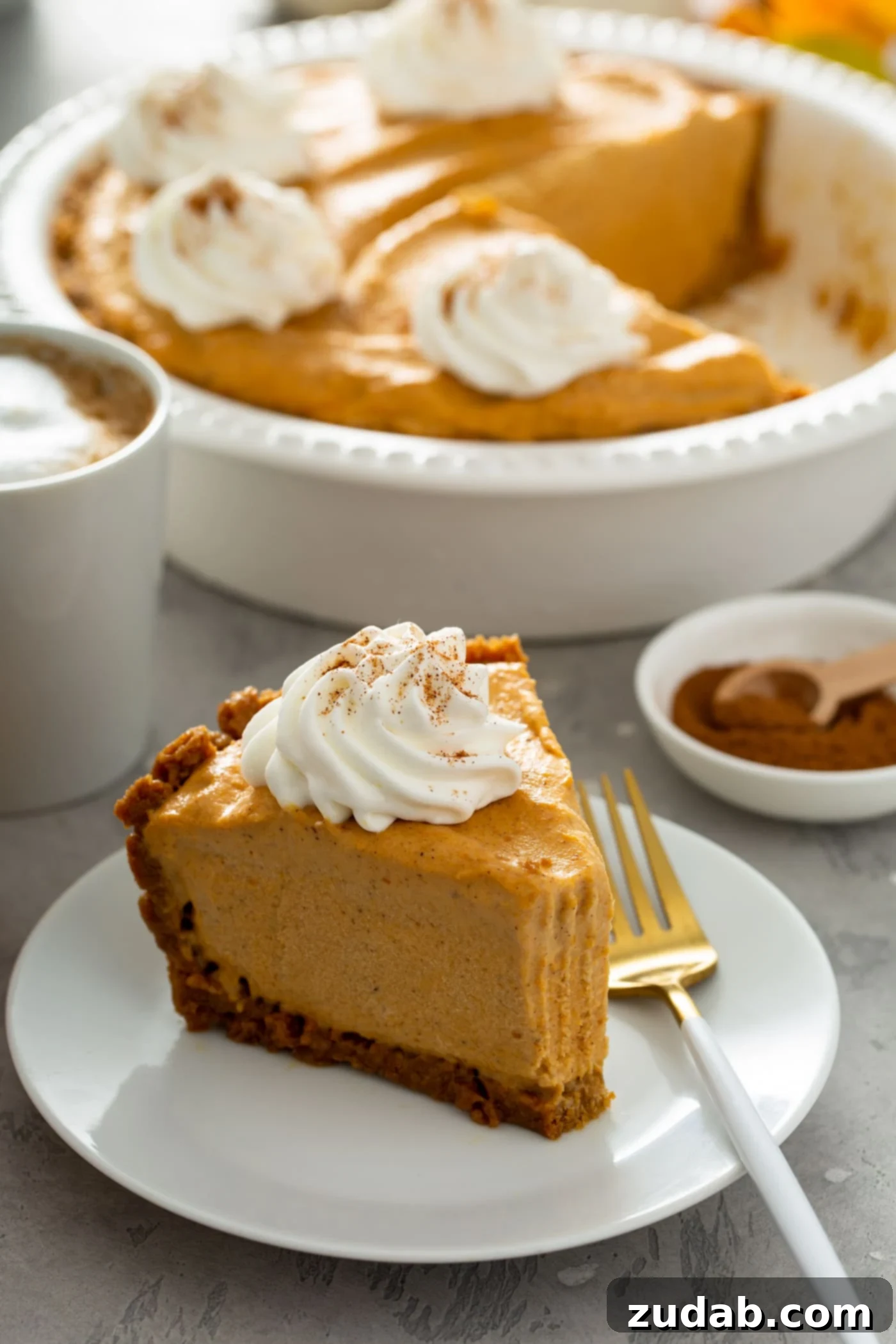 A tempting slice of pumpkin ice cream pie rests on a dessert plate, accompanied by a fork. A perfect bite has been taken from the pie, which is beautifully topped with a dollop of whipped cream sprinkled with cinnamon. In the background, a small bowl of cinnamon with a wooden spoon, a white mug of hot cocoa topped with whipped cream, and a white pie dish holding the remainder of the pumpkin ice cream pie complete a cozy and inviting scene.