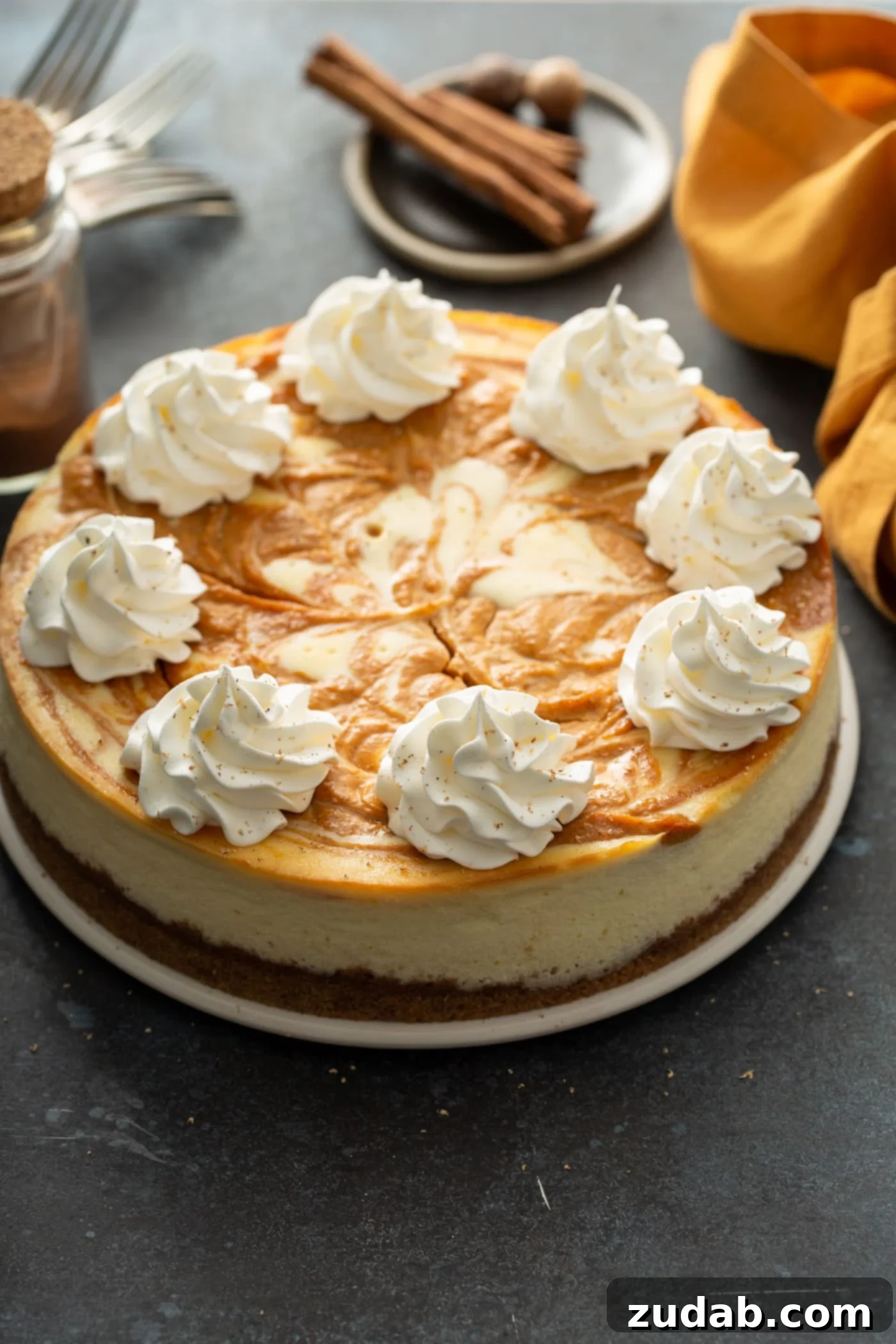 Swirled Pumpkin Cheesecake A beautiful whole swirled pumpkin cheesecake, generously topped with soft dollops of whipped cream. In the warm, festive background, there's a delicate napkin, rustic cinnamon sticks, and whole nutmeg for garnish.