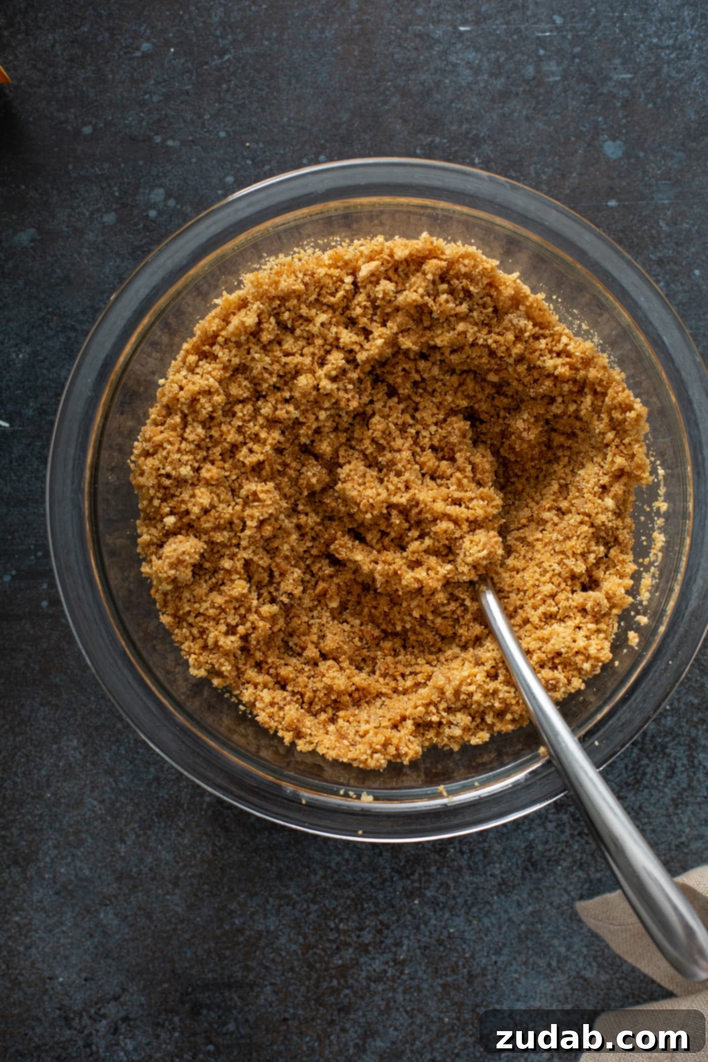 Swirled Pumpkin Cheesecake A glass bowl filled with finely crushed graham cracker crumbs, with a metal spoon resting inside, ready for the next step of mixing.