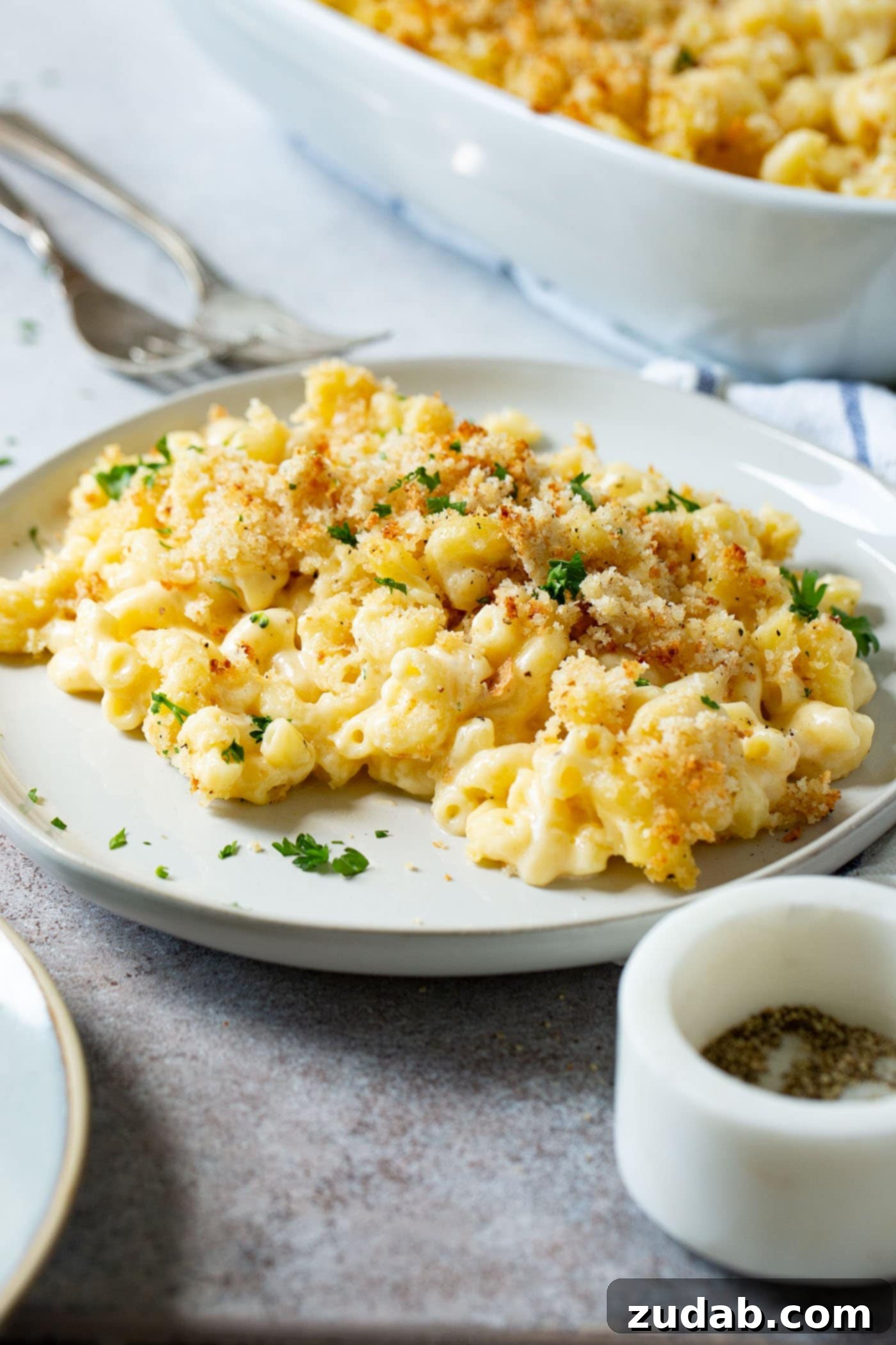 Best Macaroni and Cheese Ever A serving of the best macaroni and cheese on a dinner plate, garnished with fresh herbs. Forks are in the background, next to a pepper grinder.