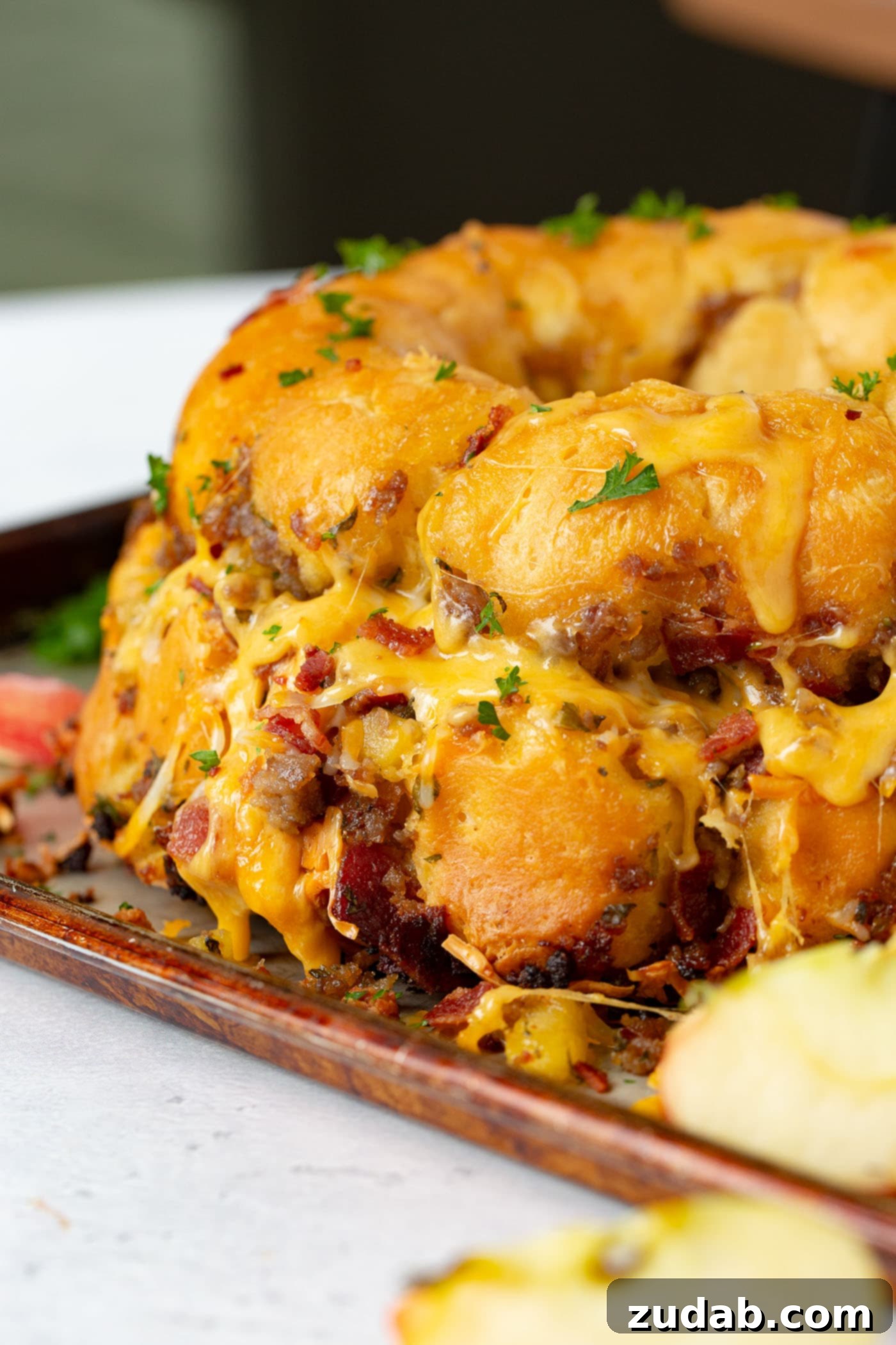 Breakfast Sausage and Apple Cheesy Pull Apart A golden-brown Breakfast Cheesy Pull Apart Bread, heavily covered in melted cheese, showcasing visible pieces of breakfast sausage, bacon, and apple. It's presented beautifully on a serving platter.