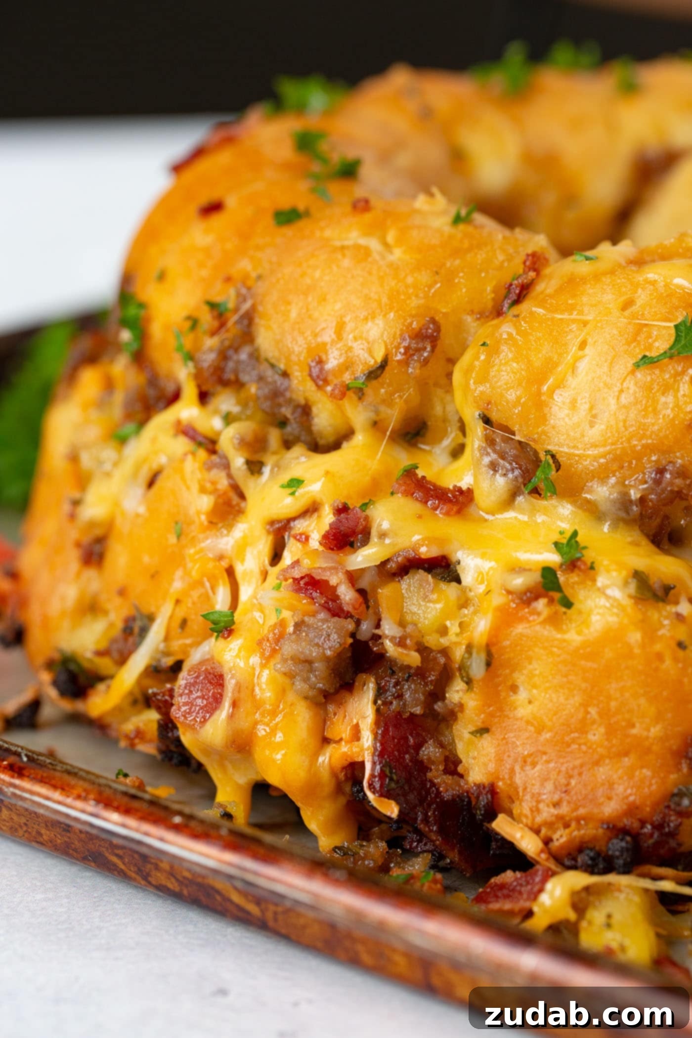 Breakfast Sausage and Apple Cheesy Pull Apart A beautiful shot of cheesy pull apart bread, still in its fluted tube pan, covered in bubbling melted cheese with visible chunks of bacon, sausage, and apple. Steam rises gently, indicating its freshly baked warmth.