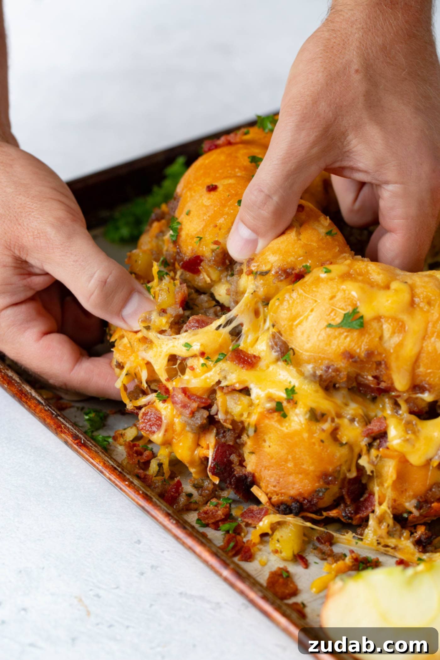 Breakfast Sausage and Apple Cheesy Pull Apart A hand reaching into a warm, cheesy Breakfast Sausage, Bacon, and Apple Pull Apart Bread, pulling a piece away. The melted cheese stretches invitingly, revealing the savory sausage, crispy bacon, and sweet apple chunks within.