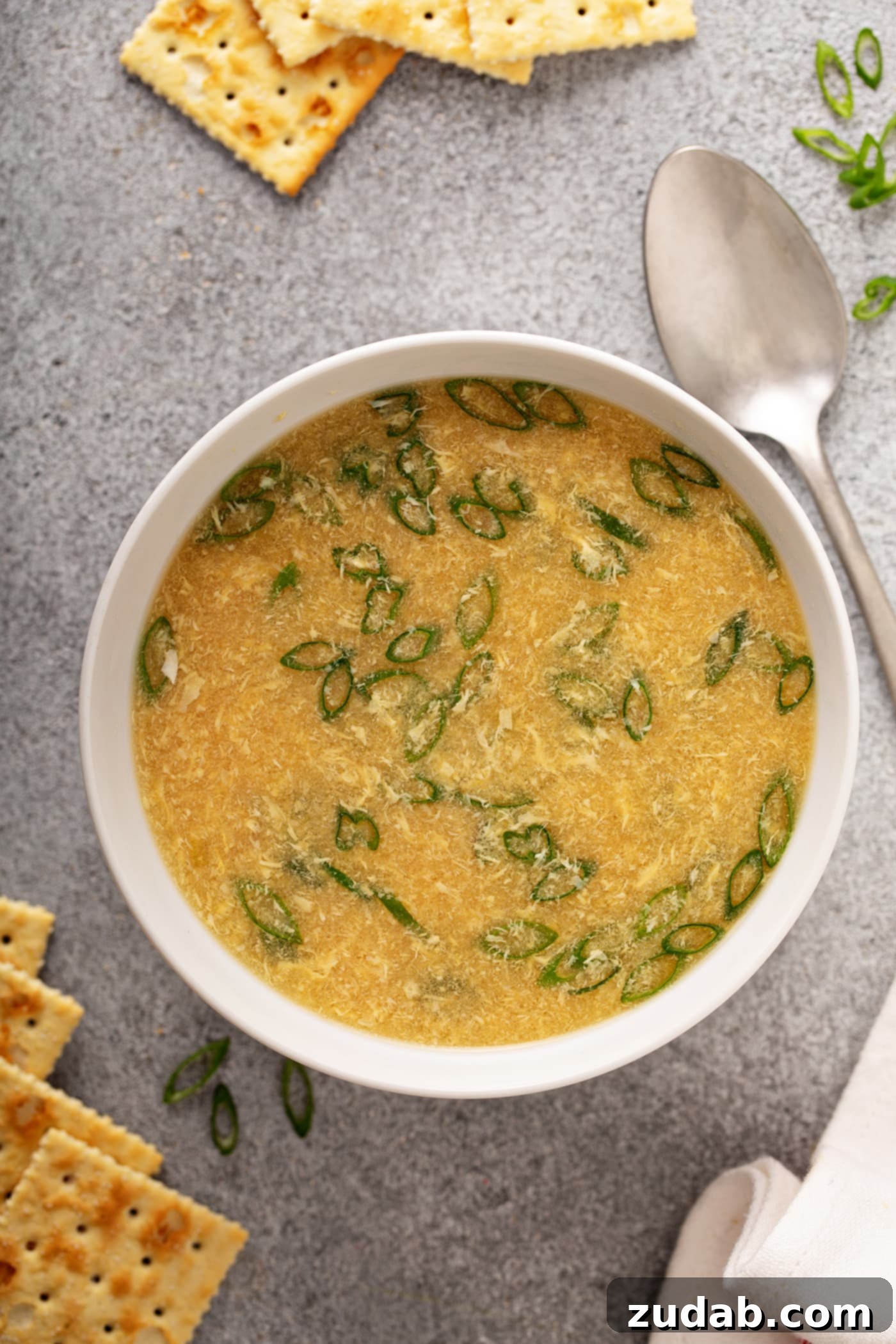 Easy Egg Drop Soup A bowl of egg drop soup with a spoon and three saltine crackers laying next to it. You can see bits of egg and sliced green peppers in the soup.