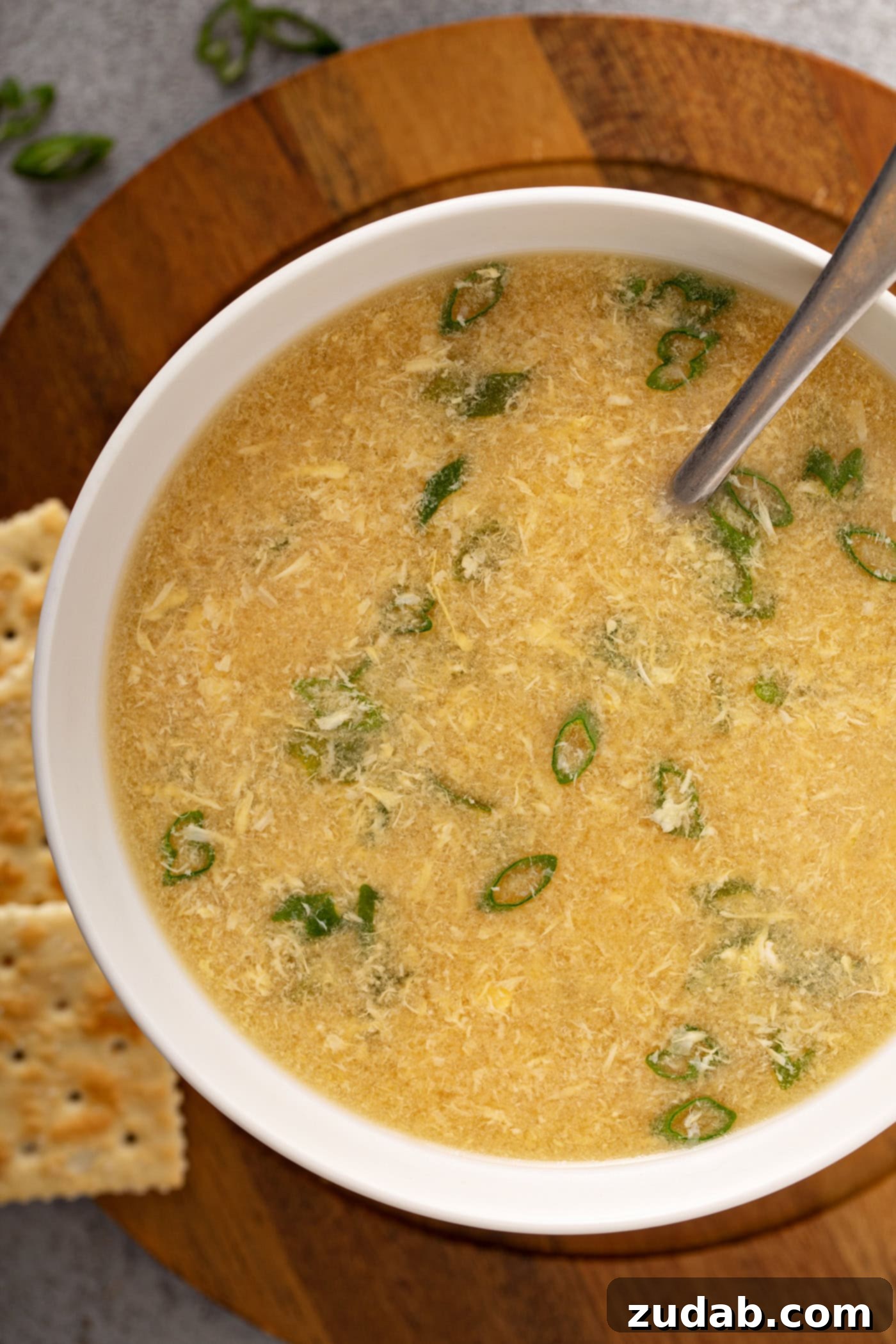 Easy Egg Drop Soup A close-up of a bowl of egg drop soup with sliced onions on top and saltine crackers in the background.