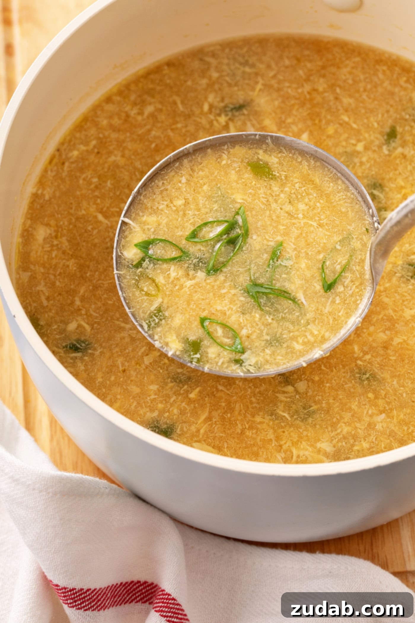 Easy Egg Drop Soup A ladle of egg drop soup above a bowl containing egg drop soup showing bits of egg and sliced green onions.