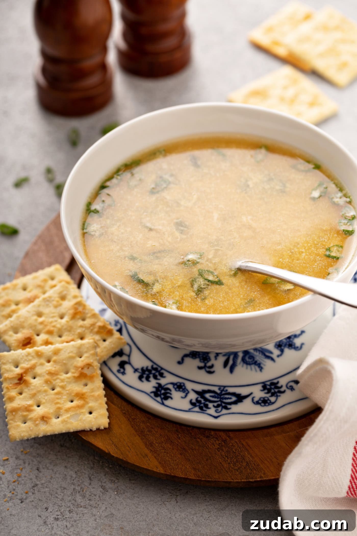 Easy Egg Drop Soup A saucer with a bowl of egg drop soup sitting on top. there is a spoon in the soup and you can see slices of green onion in the soup. Three saltine crackers are on the saucer beside the soup.