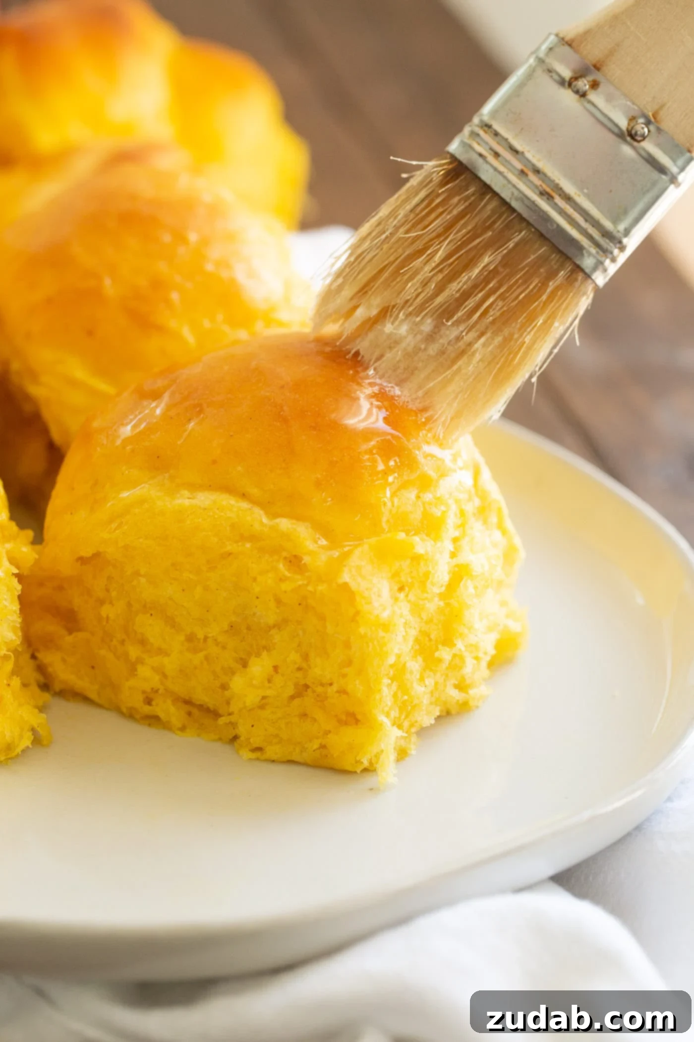 Pillowy Pumpkin Dinner Rolls 5 A freshly baked pumpkin dinner roll being brushed with melted butter for a glistening finish