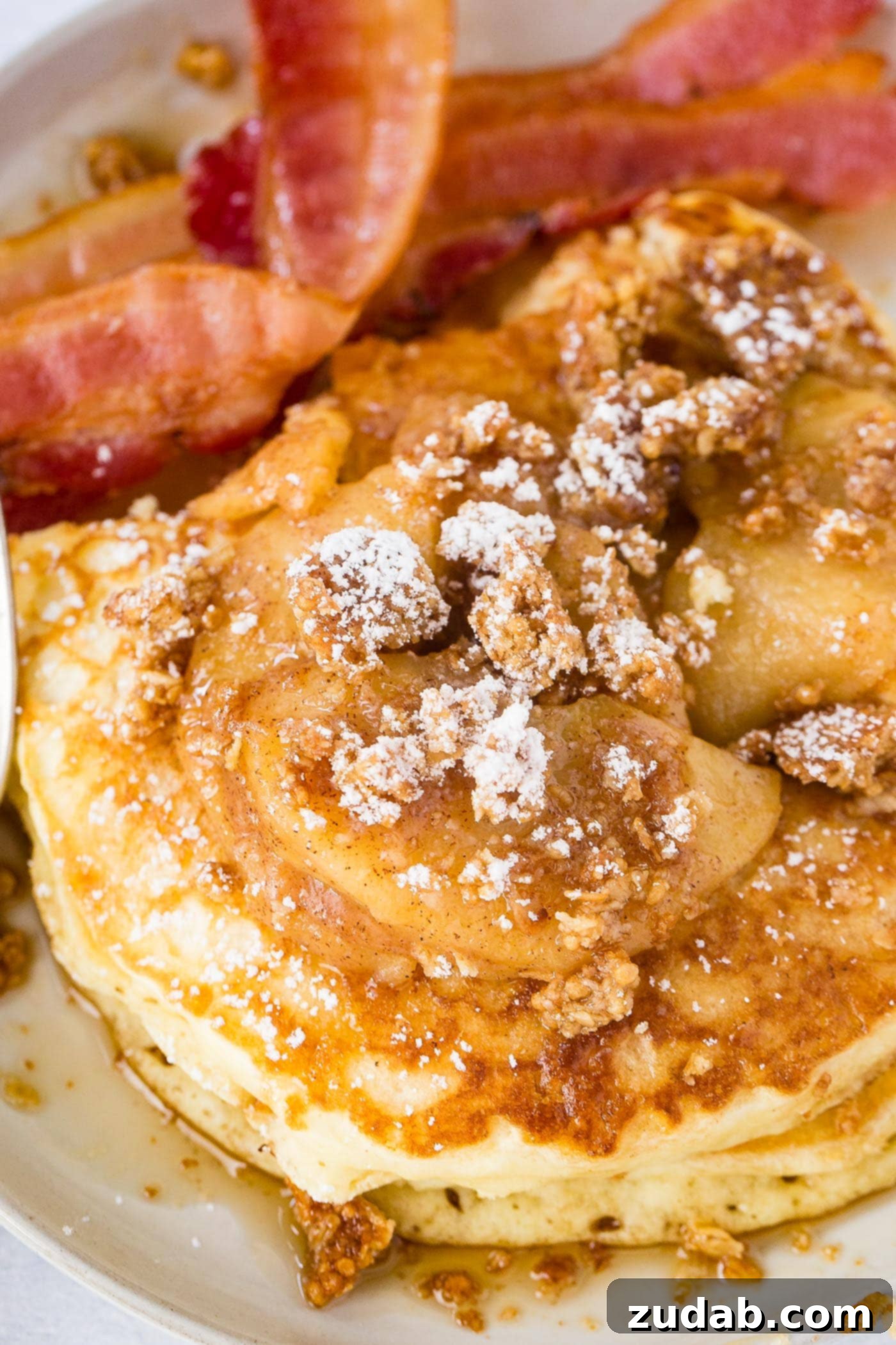Disneyland-Inspired Apple Granola Pancakes 2 a plate stacked with fluffy pancakes topped with roasted apples, crumbled granola, powdered sugar and a drizzle of syrup with a side of bacon.