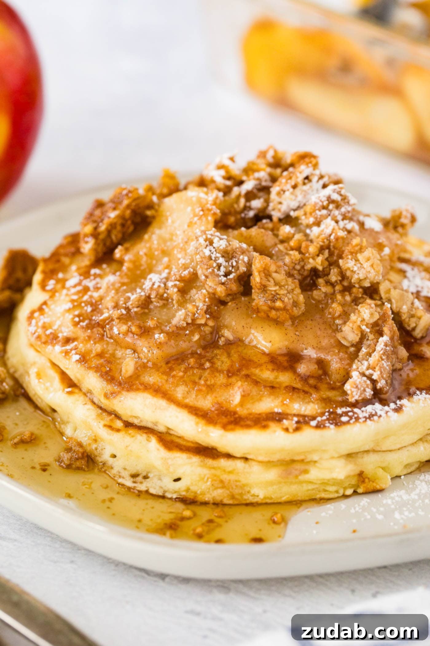 Disneyland-Inspired Apple Granola Pancakes 4 a plate stacked with fluffy pancakes topped with roasted apples, crumbled granola, powdered sugar and a drizzle of syrup with a side of bacon.