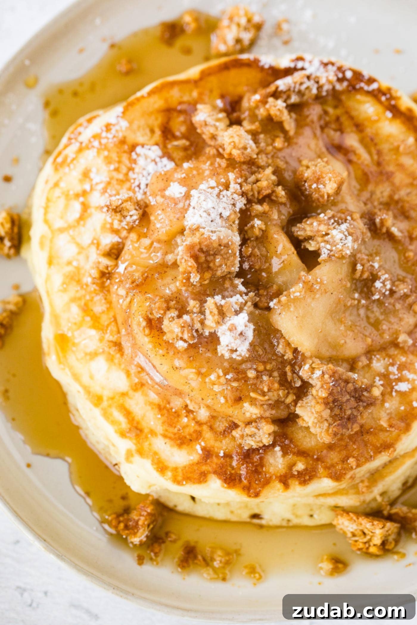 Disneyland-Inspired Apple Granola Pancakes 6 a plate stacked with fluffy pancakes topped with roasted apples, crumbled granola, powdered sugar and a drizzle of syrup with a side of bacon.