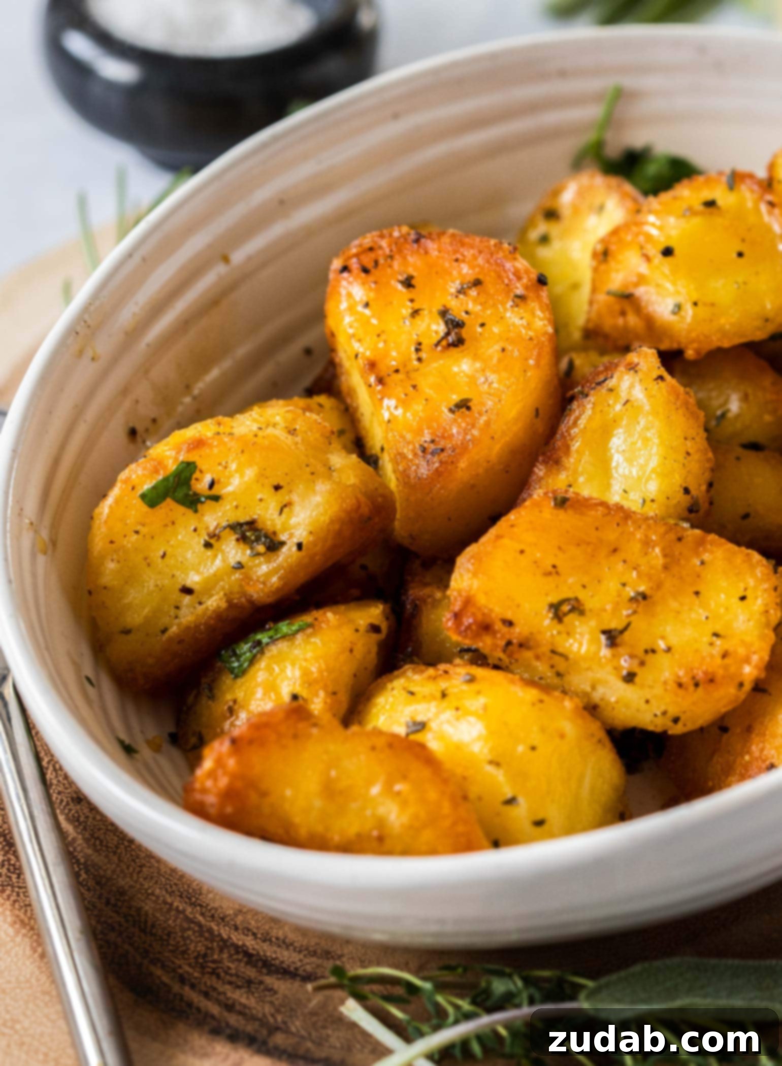 Crispy Garlic-Herb Roasted Potatoes 3 A cream coloured bowl overflowing with golden-brown, glistening herby roasted potatoes, garnished with fresh herbs. The bowl sits on a light wooden board, ready to be served.