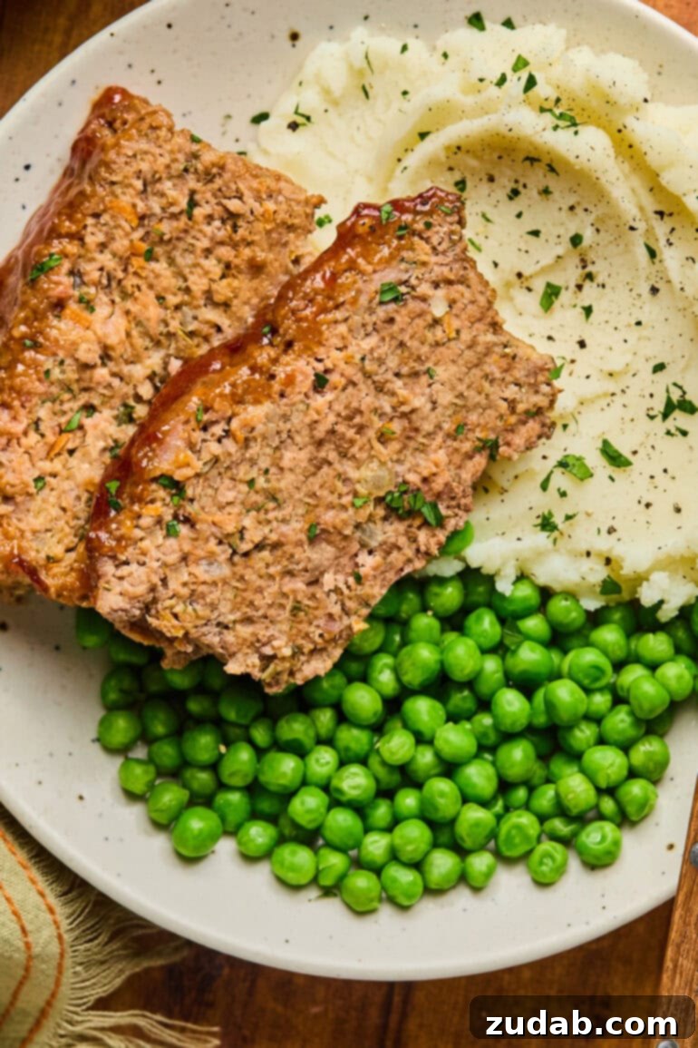 Our Favorite Classic Meatloaf 2 A perfectly cooked plate of classic meatloaf slices served with creamy mashed potatoes and vibrant green peas, a quintessential comfort meal.