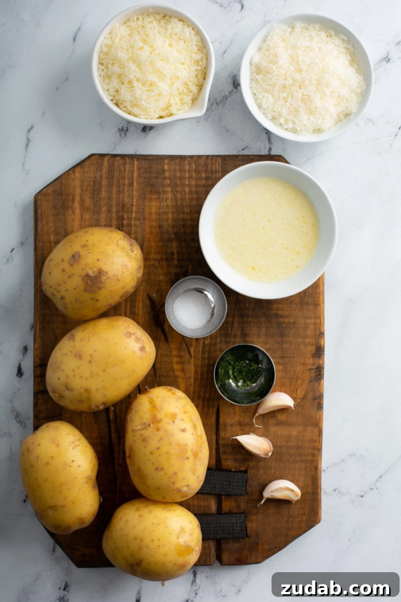 A wooden cutting board displaying fresh ingredients for Gruyere potatoes: four Yukon Gold potatoes, three cloves of garlic, coarse sea salt, black pepper, a bowl of shredded Gruyere and Parmesan cheese, and a small bowl of melted butter.