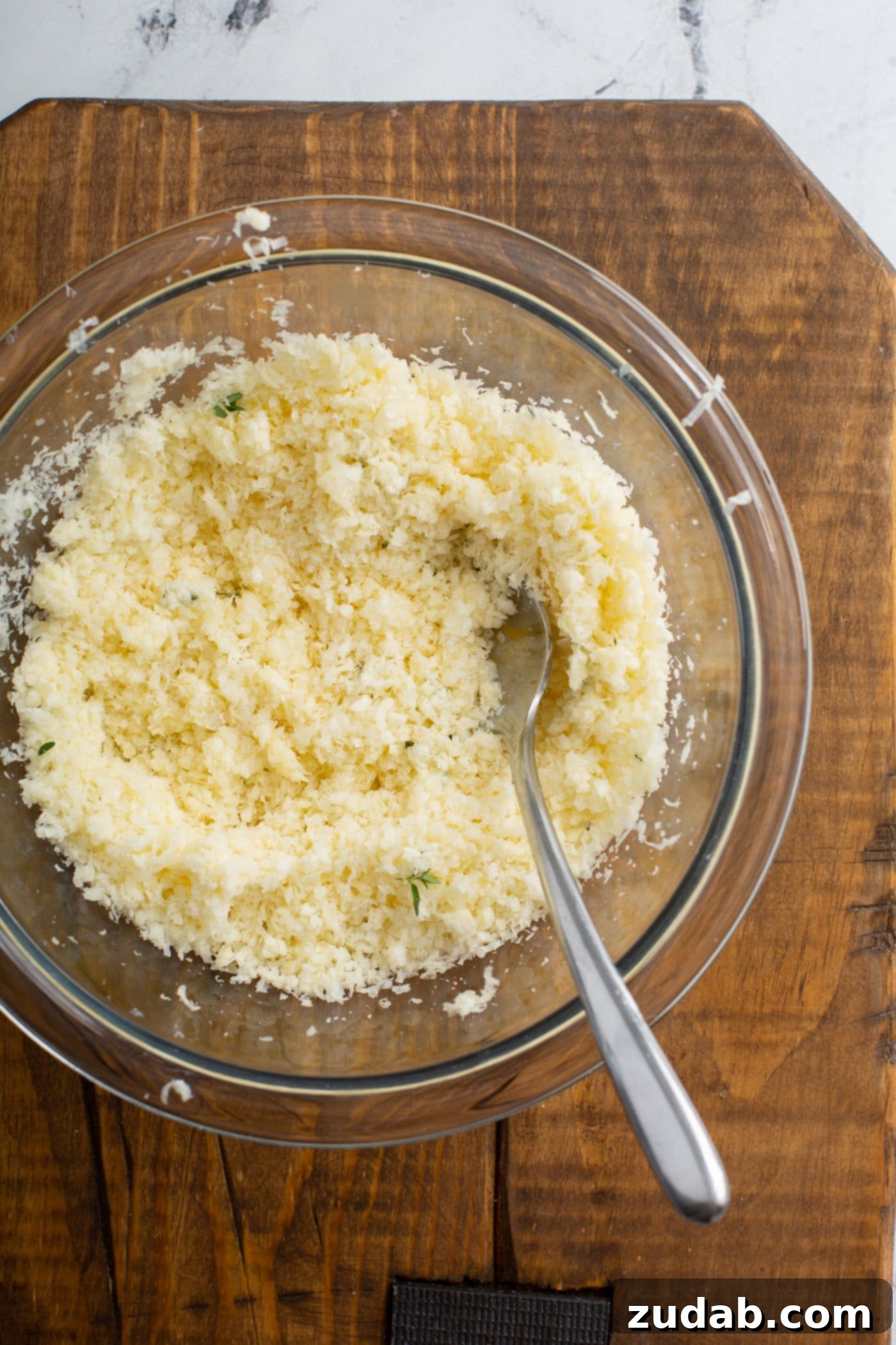 A bowl filled with a fragrant mixture of finely shredded Gruyere and Parmesan cheeses, minced garlic, fresh thyme, and coarse sea salt, with a spoon resting inside.