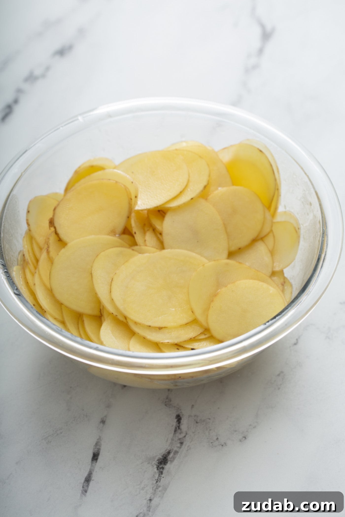 A clear glass bowl filled with thinly sliced Yukon Gold potatoes, shimmering with a coating of melted butter, ready for layering.