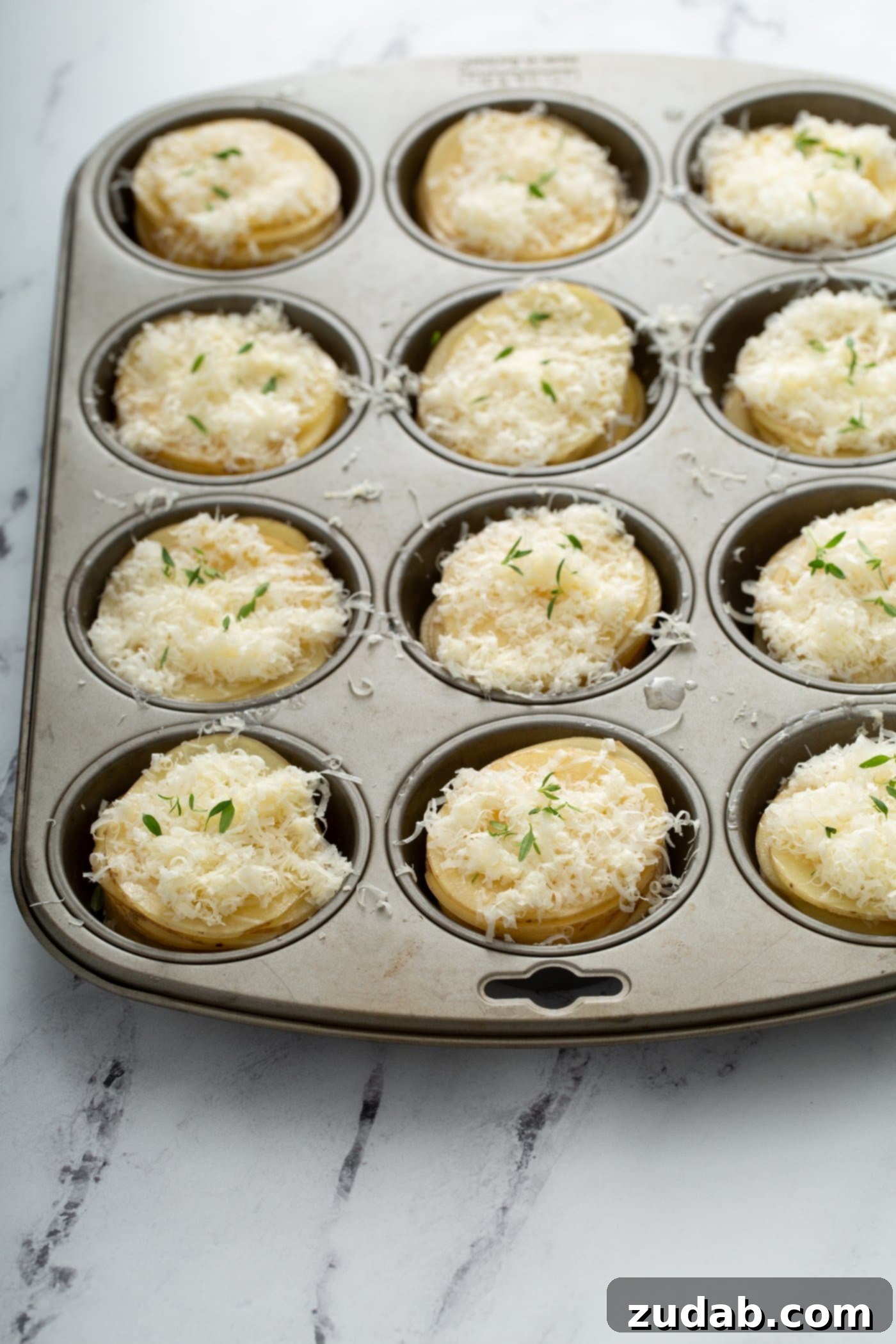 A muffin tin with individual cups filled with artfully layered thin potato slices and generous portions of shredded Gruyere and Parmesan cheeses, ready for baking.