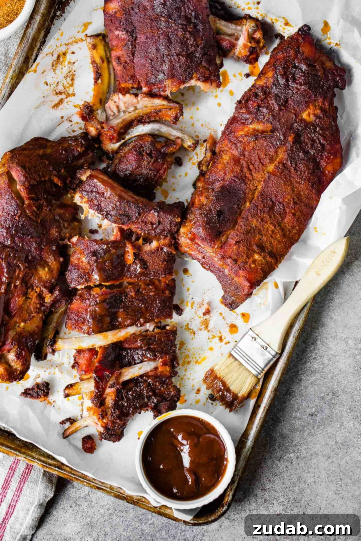 A photo of 3 racks of smoked ribs, two of them sliced into servings and one whole with a sauce brush and BBQ sauce on the side.