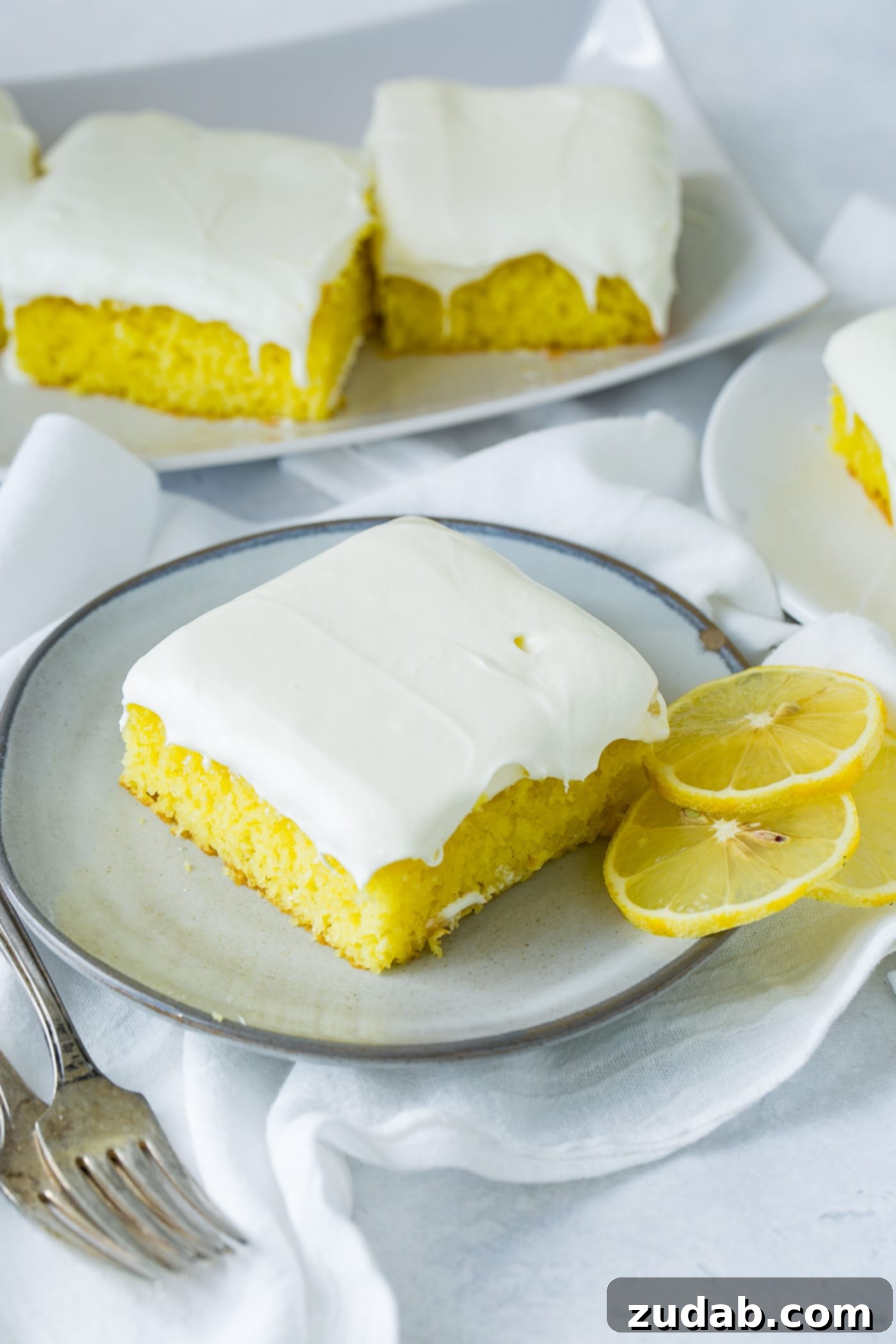 A slice of frosted lemon jello cake with two round slices of lemon on a serving plate. There is a plate containing more slices in the background and white linen napkins under the plates.