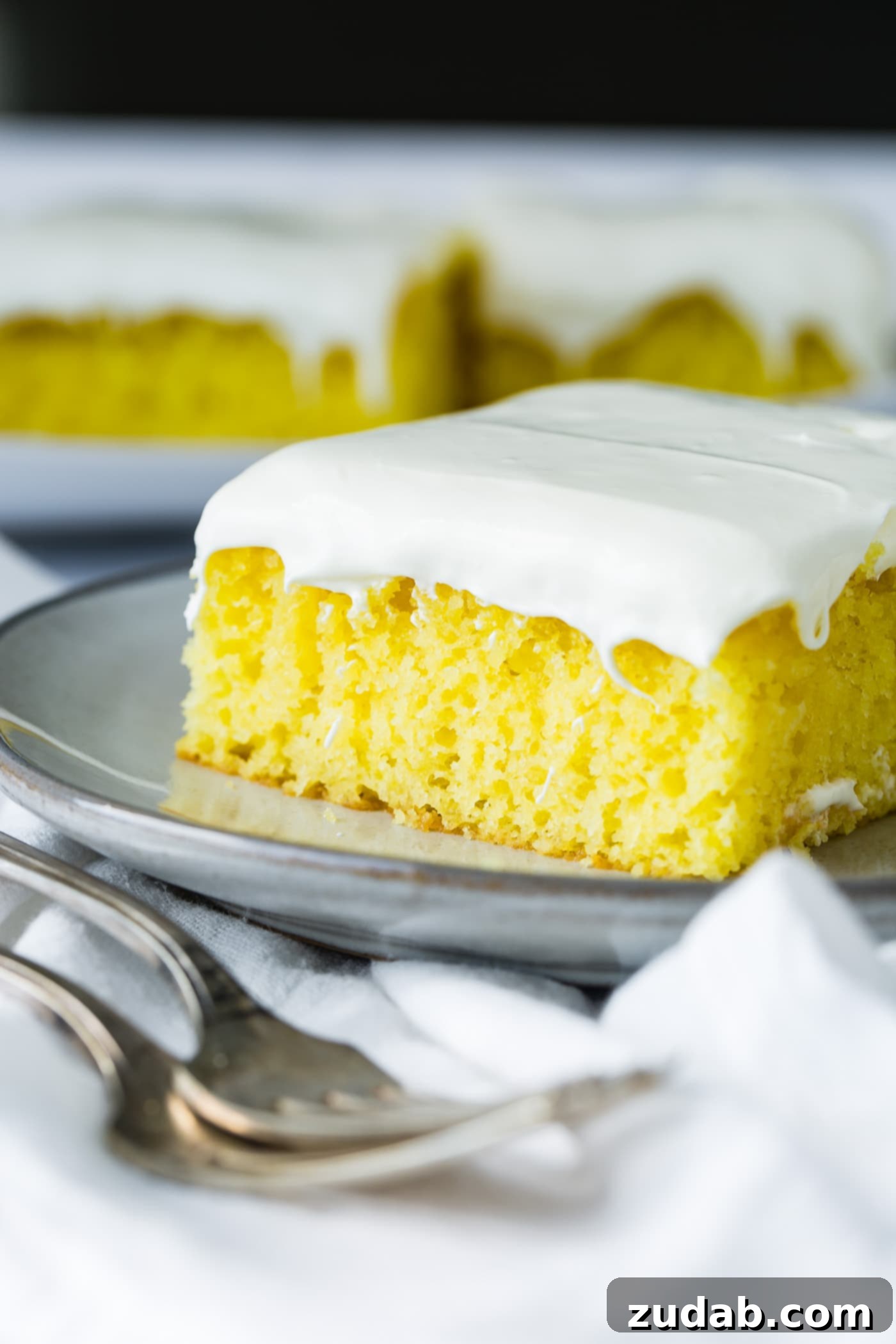 A slice of frosted lemon jello cake on a serving plate with a couple of slices on another plate in the background. There are two forks resting on a white linen napkin next to the plate of cake.