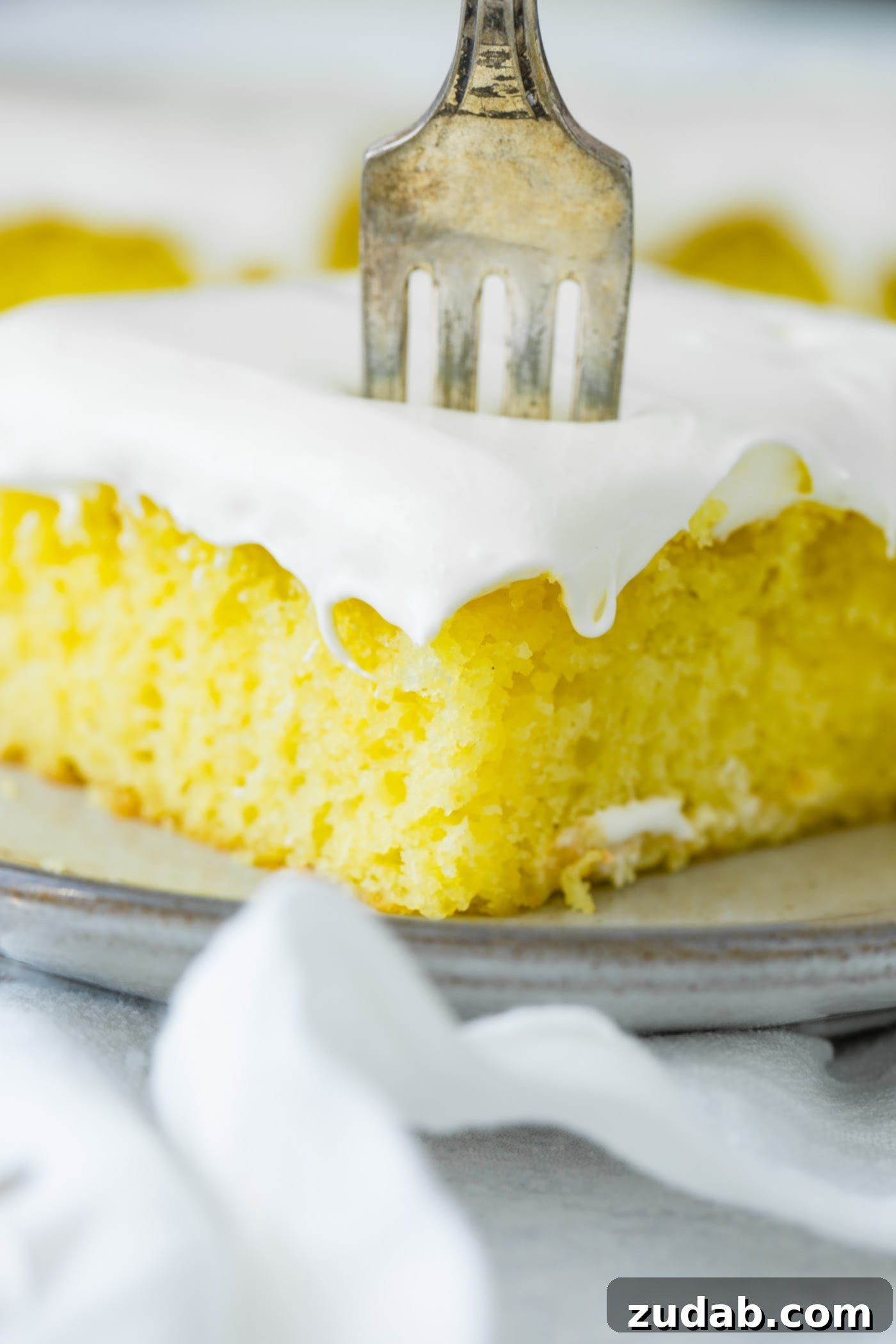 A slice of frosted lemon jello cake with a fork sticking into the top. It is on a serving plate which is sitting on a white linen napkin.