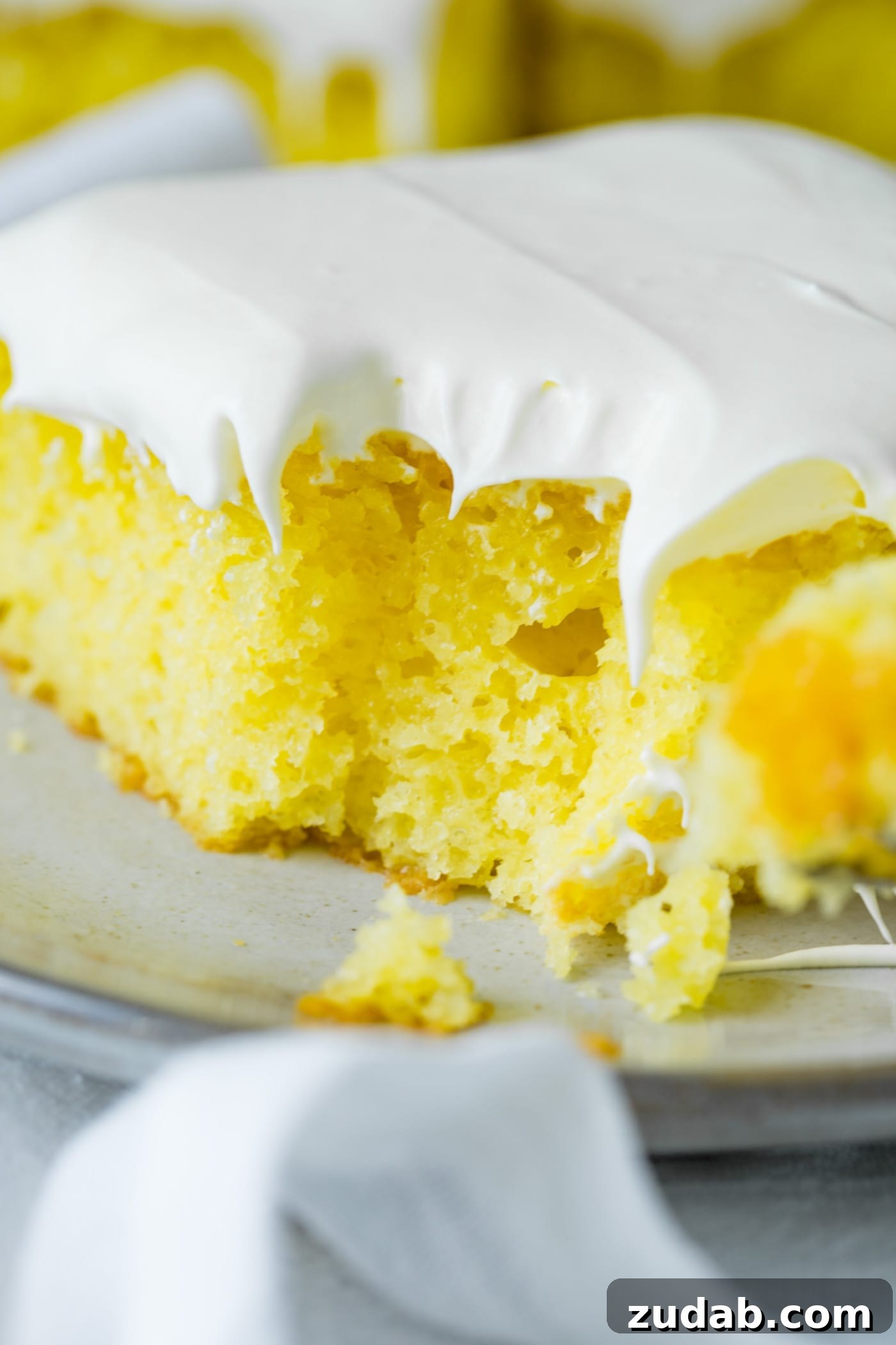 A slice of bright yellow lemon jello cake with fluffy white frosting on top. A bite has been taken from the corner of the cake.