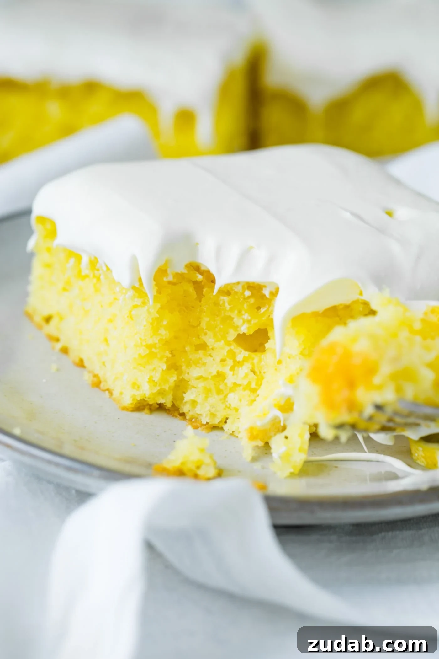 A slice of yellow lemon jello cake with a bite taken from the corner. It is topped with a fluffy white frosting and rests on a serving plate on top of a white linen napkin.
