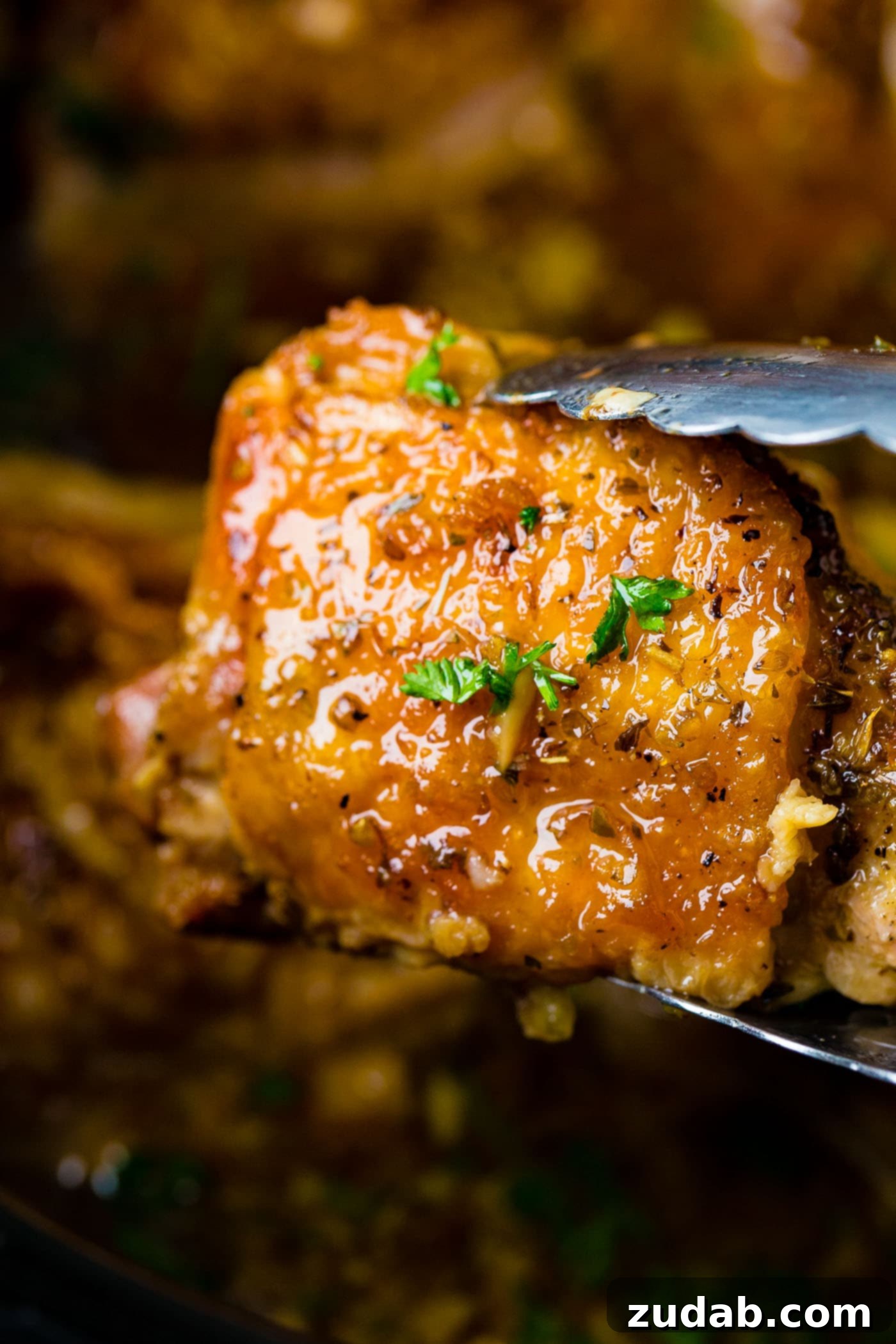A pair of metal tongs holding up a perfectly cooked, golden-brown chicken thigh, showcasing the succulence of our Slow Cooker Garlic Chicken.