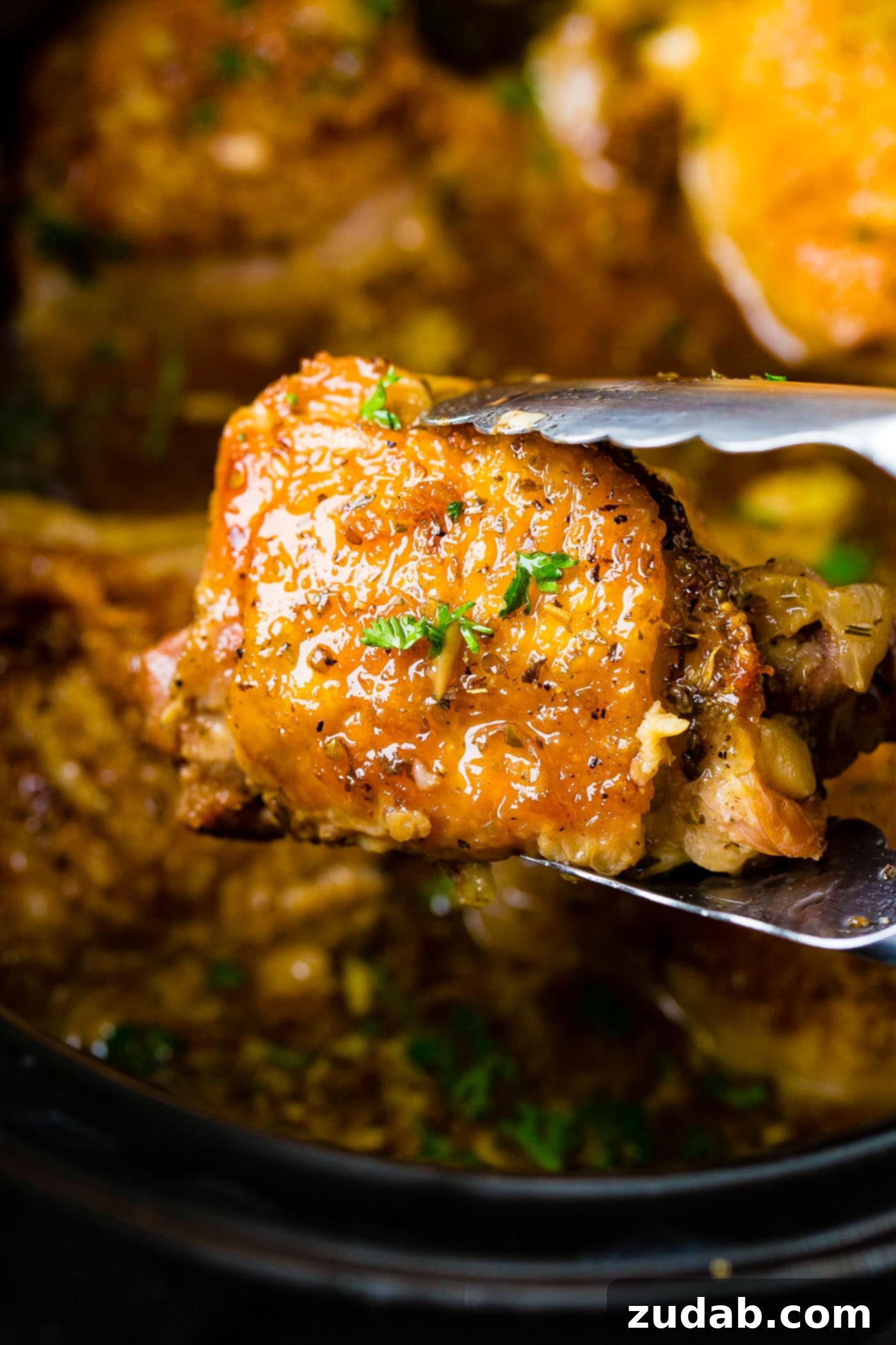 A pair of metal tongs holding up a perfectly cooked, golden-brown chicken thigh from our Slow Cooker Garlic Chicken recipe, emphasizing its tender texture.