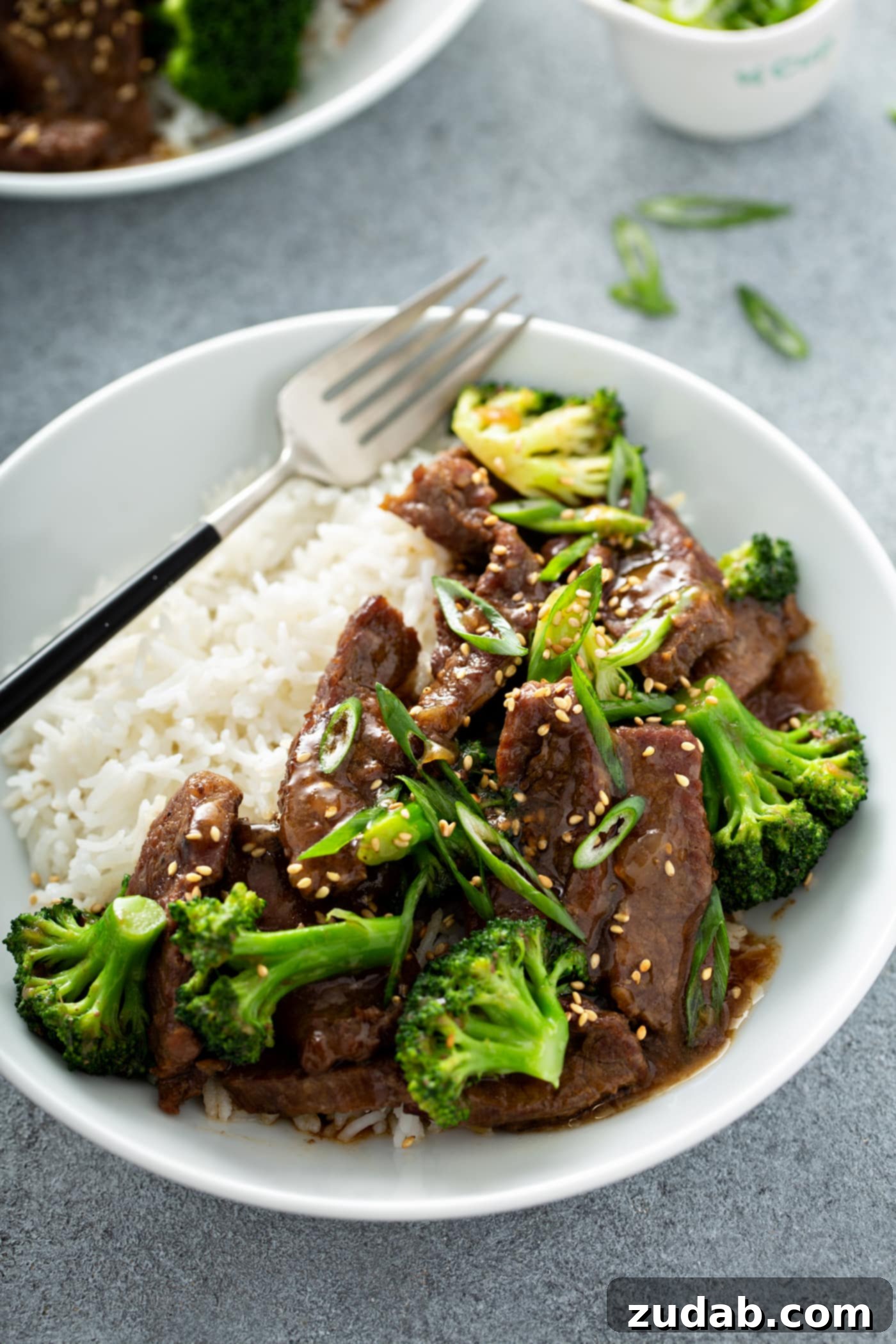 Instant Pot Beef and Broccoli A photo of beef and broccoli on a white plate next to a serving of white rice and garnished with sliced green onions and toasted sesame seeds