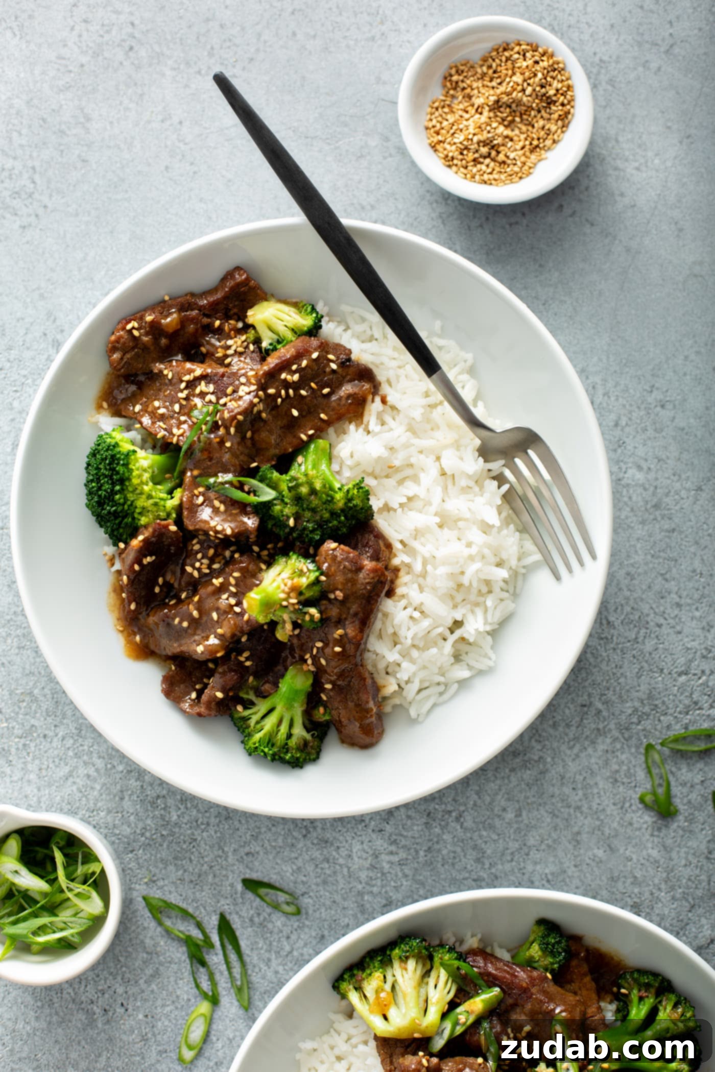 Instant Pot Beef and Broccoli A photo of a white bowl with a serving of beef and broccoli in it along side some white rice. There is also a small dish of toasted sesame seeds next to the bowl.