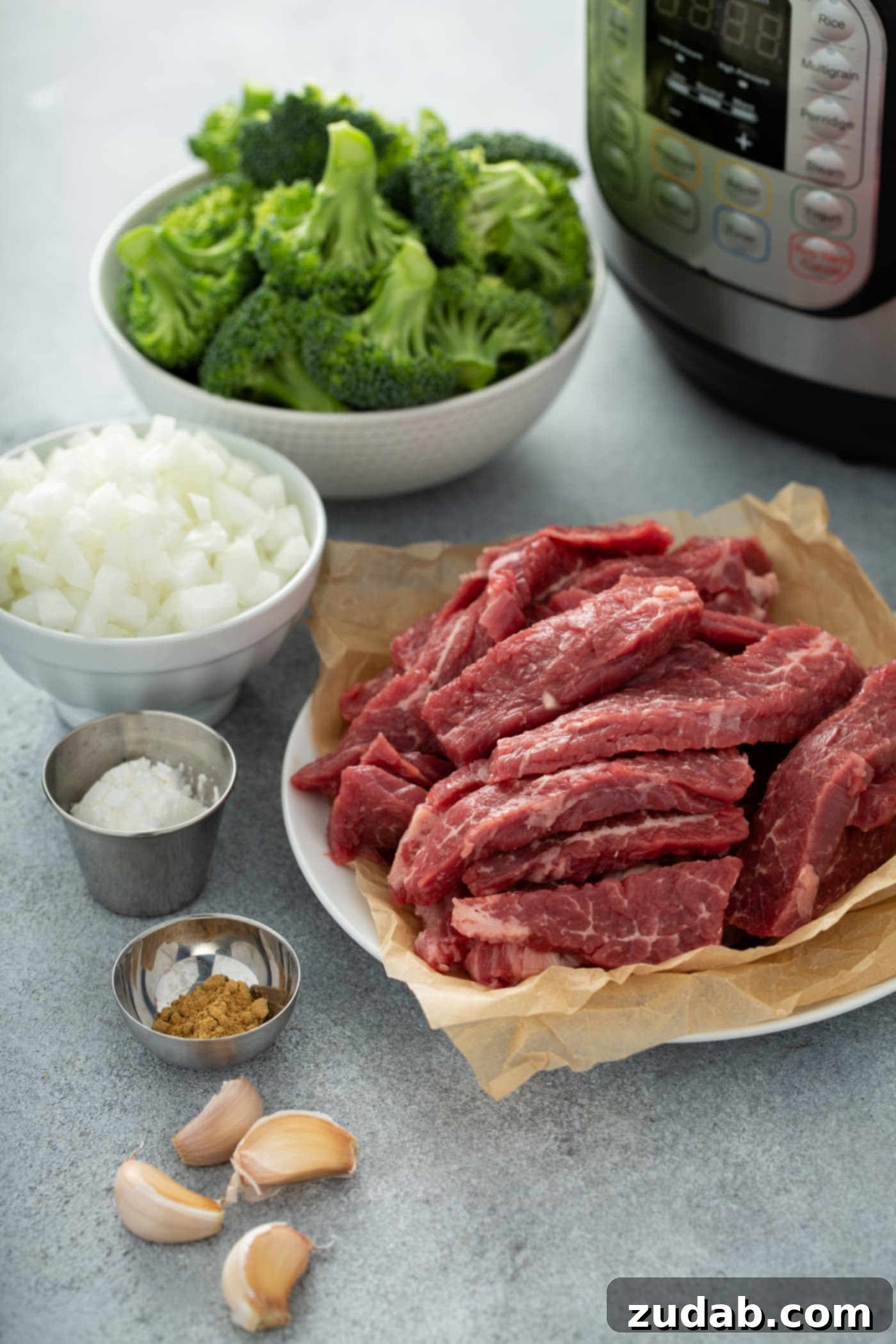 Instant Pot Beef and Broccoli A photo of the ingredients needed to make beef and broccoli with an Instant Pot in the background. The ingredients are in separate bowls, including broccoli florets, chopped onion, sliced raw flank steak, cornstarch, ground ginger and garlic cloves.